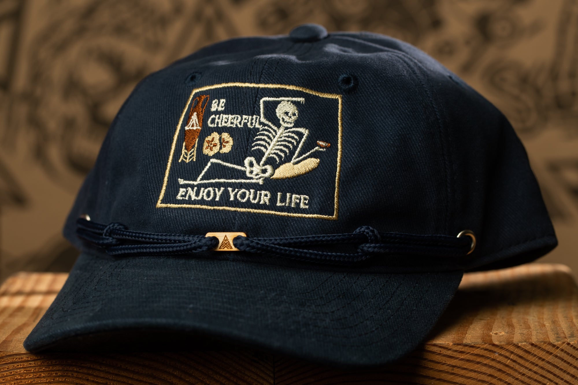 A navy blue baseball cap with a white and gold embroidered patch on the front, featuring a skeleton and the text "Be cheerful, enjoy your life".
