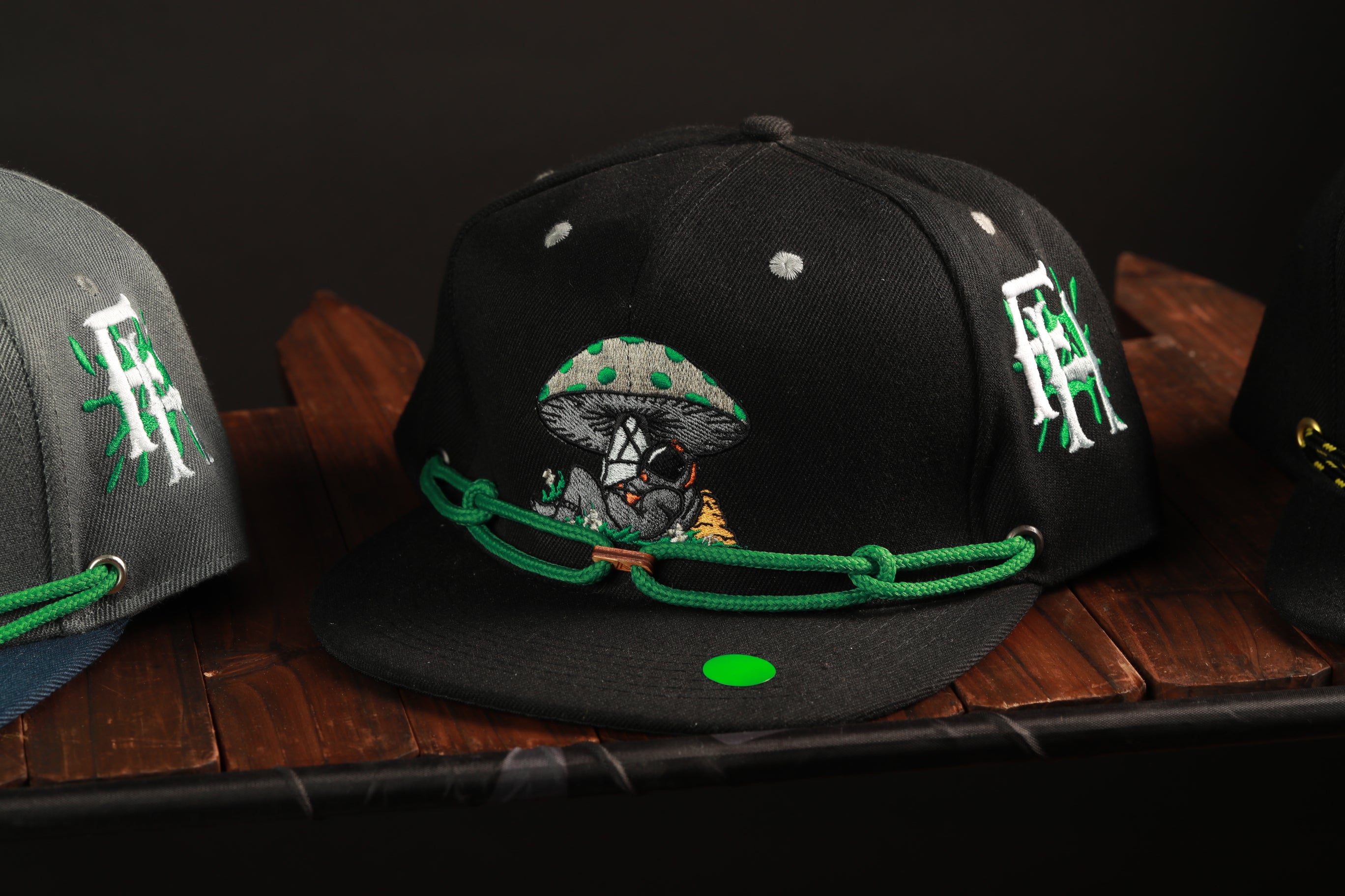 A black baseball cap with a mushroom design and green strings is displayed on a wooden surface.
