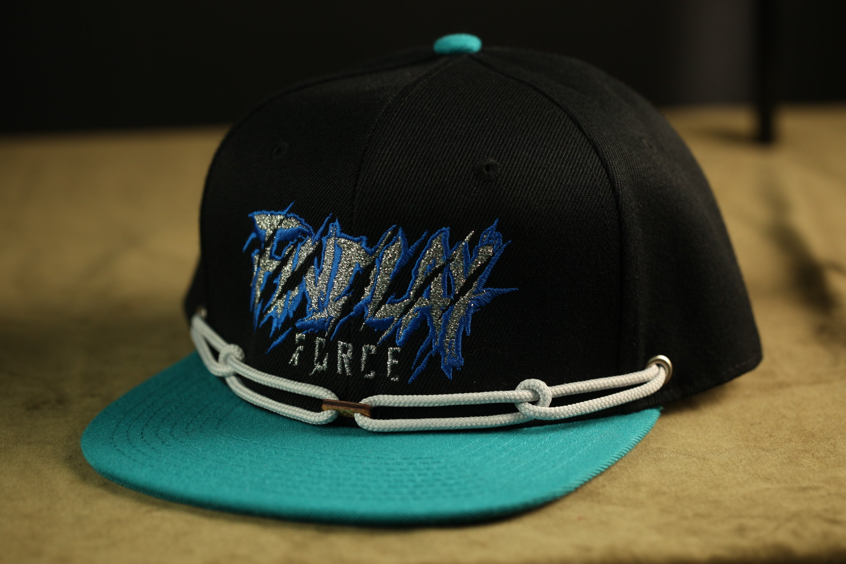 A black baseball cap with a teal brim and a white chain strap, featuring a blue and white graphic design on the front.