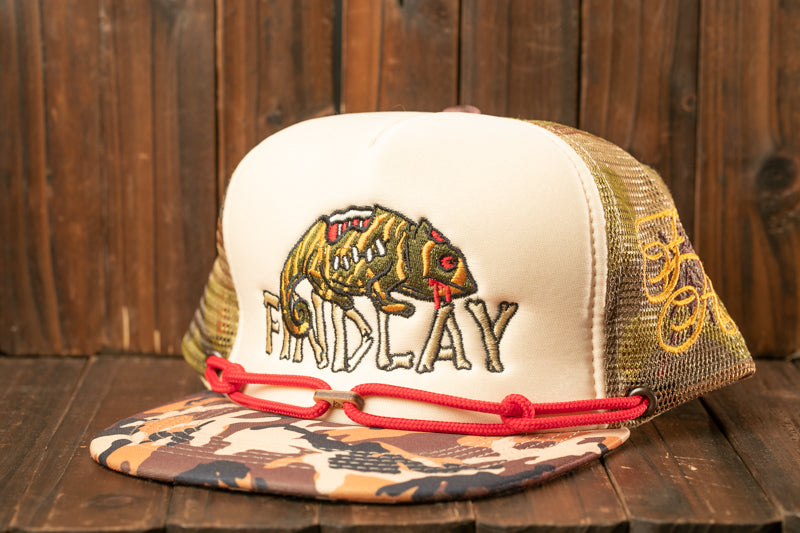 A camouflage baseball cap with a printed design of a chameleon on it, sitting on a wooden surface.