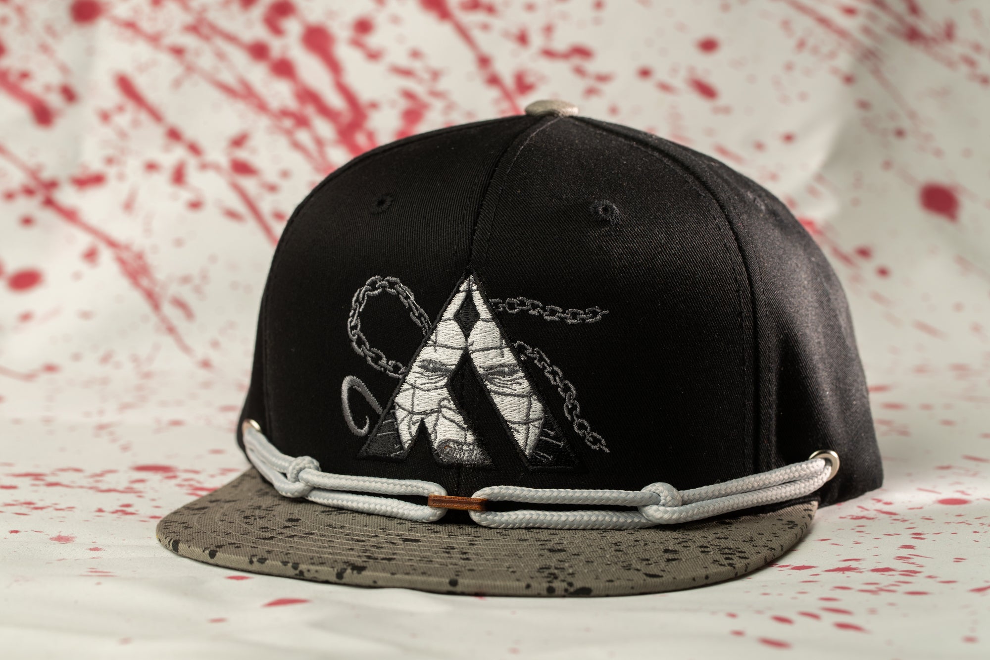 A black baseball cap with a chain design on the front, sitting on a speckled surface with red splatters.
