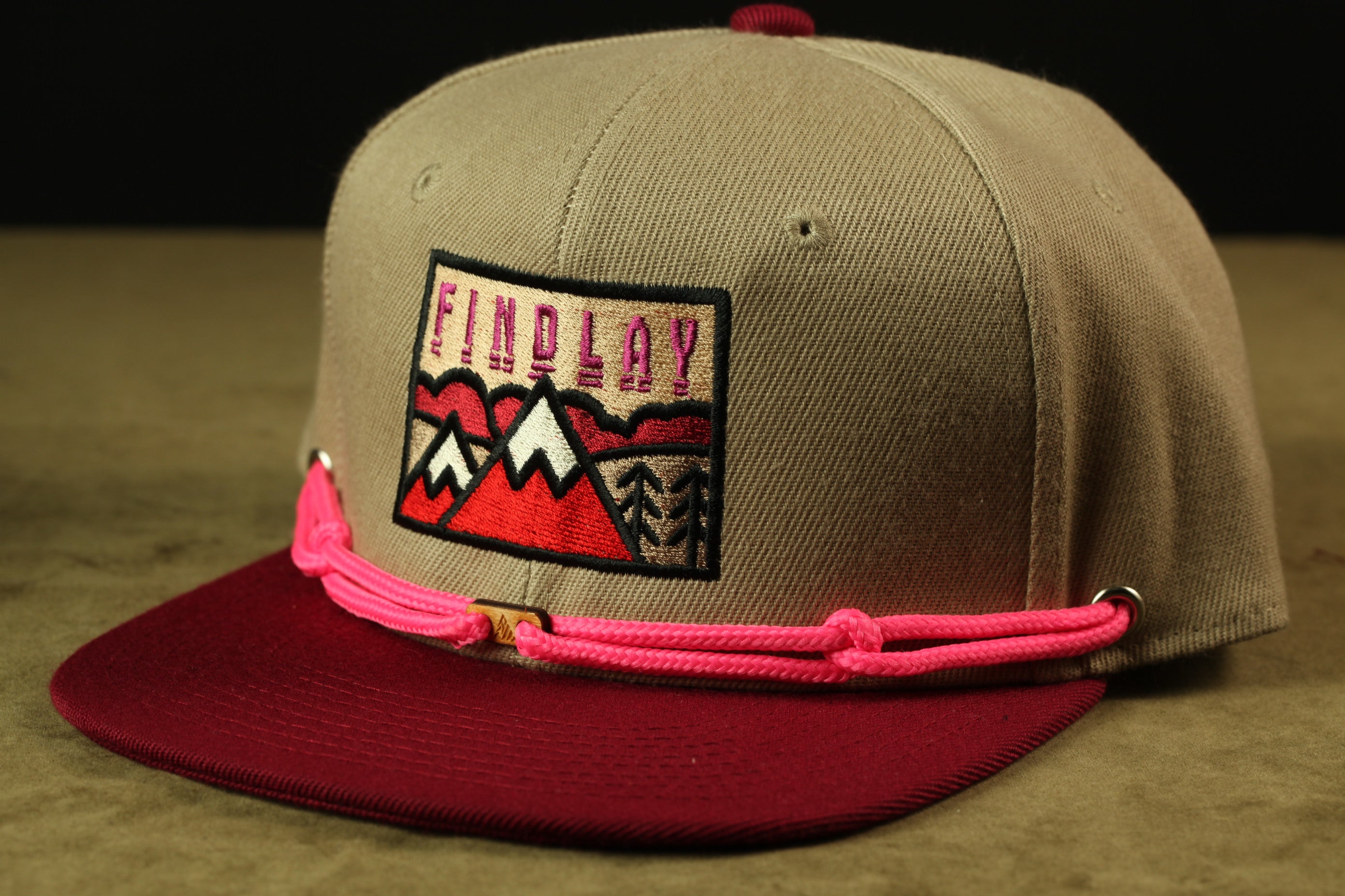 A tan baseball cap with a maroon brim and a pink braided strap, featuring a graphic design on the front.