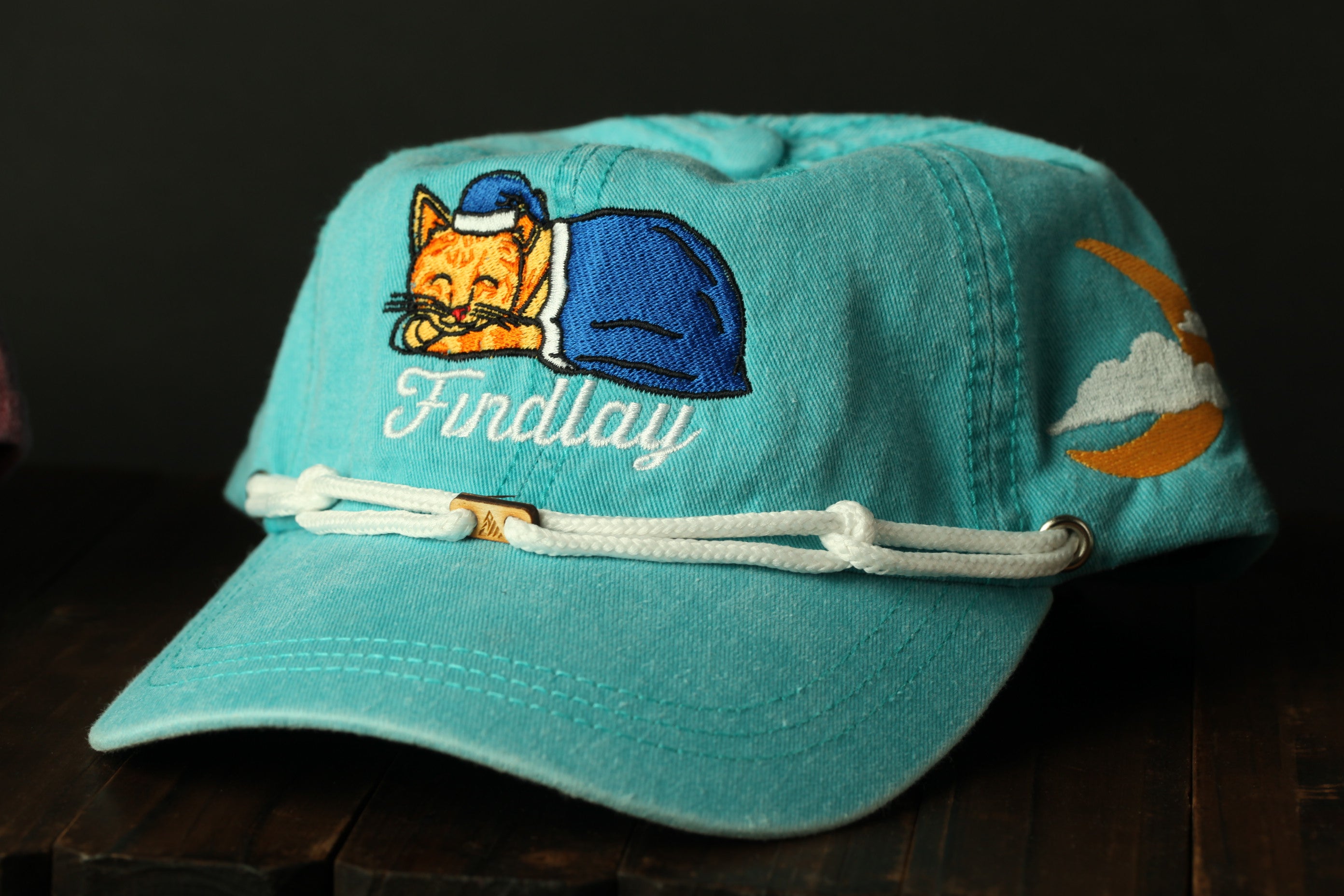 A turquoise baseball cap with a cat embroidered on it, featuring a blue blanket and a crescent moon.