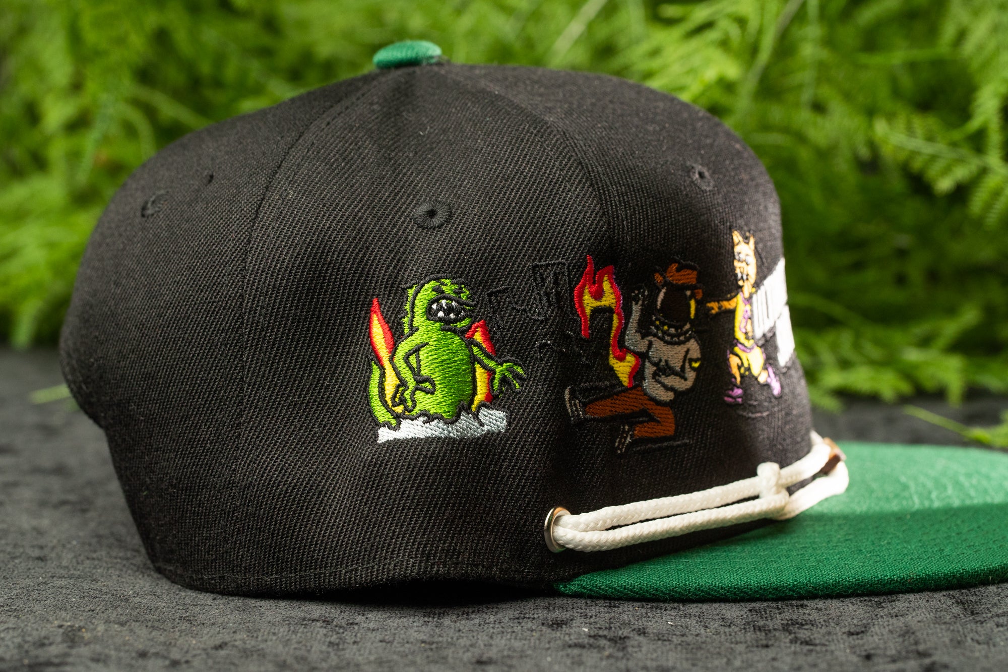 A black baseball cap with colorful cartoon-style characters and designs on it, including a green alien, a green dinosaur, and a green cat.