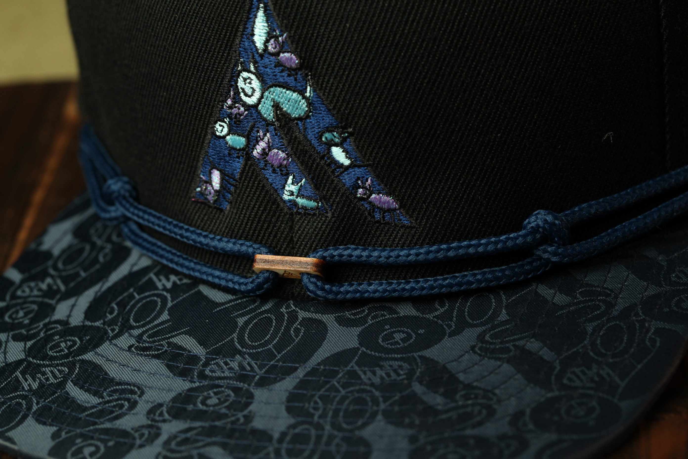 A close-up view of a black baseball cap with a blue and purple embroidered logo on the front.