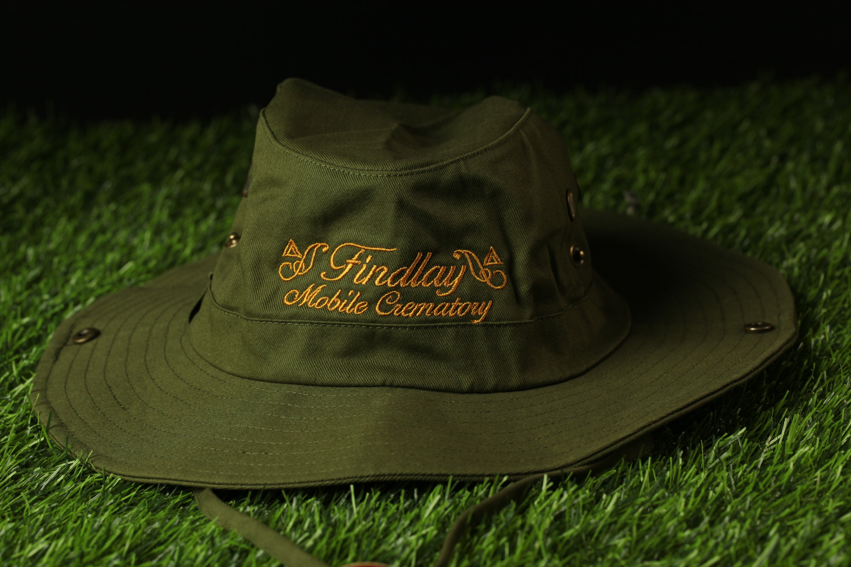 A green bucket hat with the text "Findlay Mobile Cematory" printed on it, placed on a grassy surface.