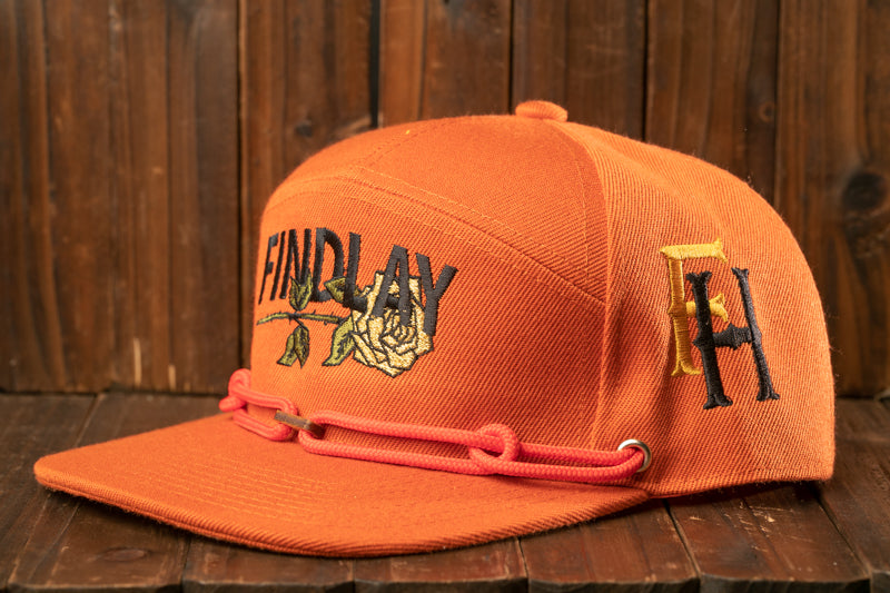 An orange baseball cap with the text "FIND LAY" printed on it, sitting on a wooden surface.