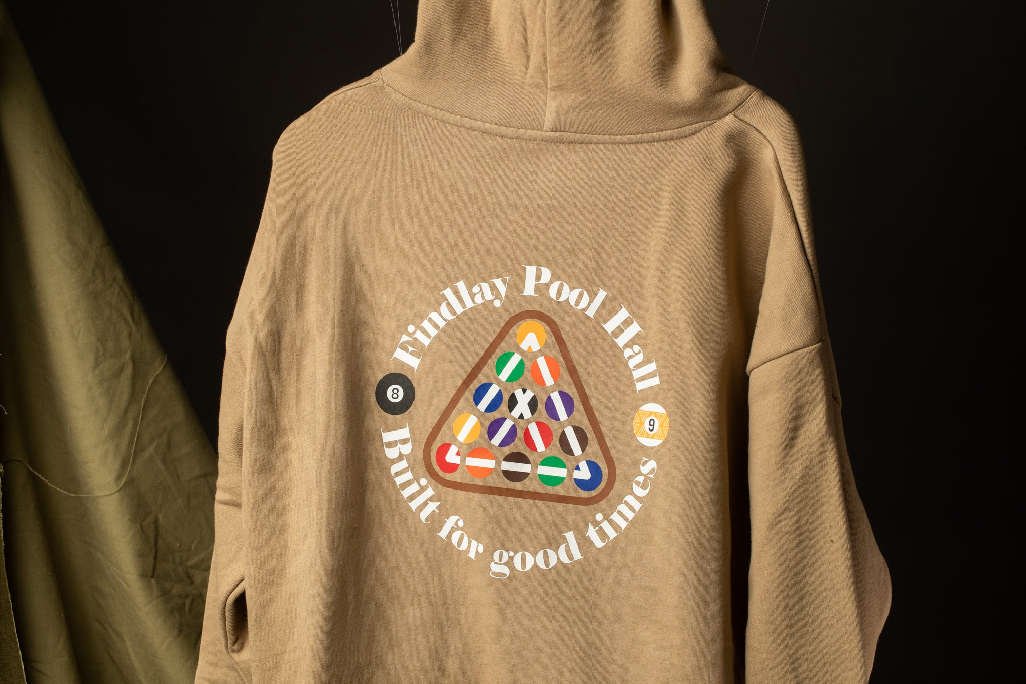 A beige hoodie with a graphic design on the back, featuring a pool table with colorful balls and the text "Findlay Pool Hall Built for good times".