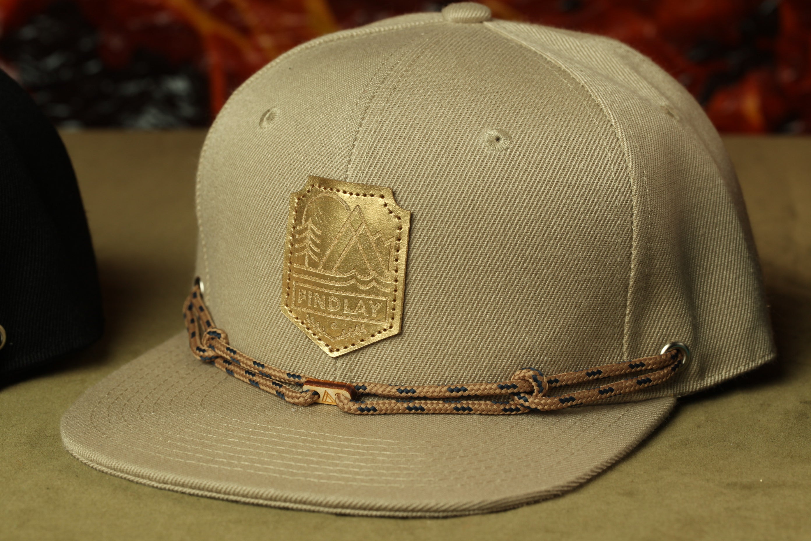 A tan baseball cap with a gold-colored logo and a brown braided strap.