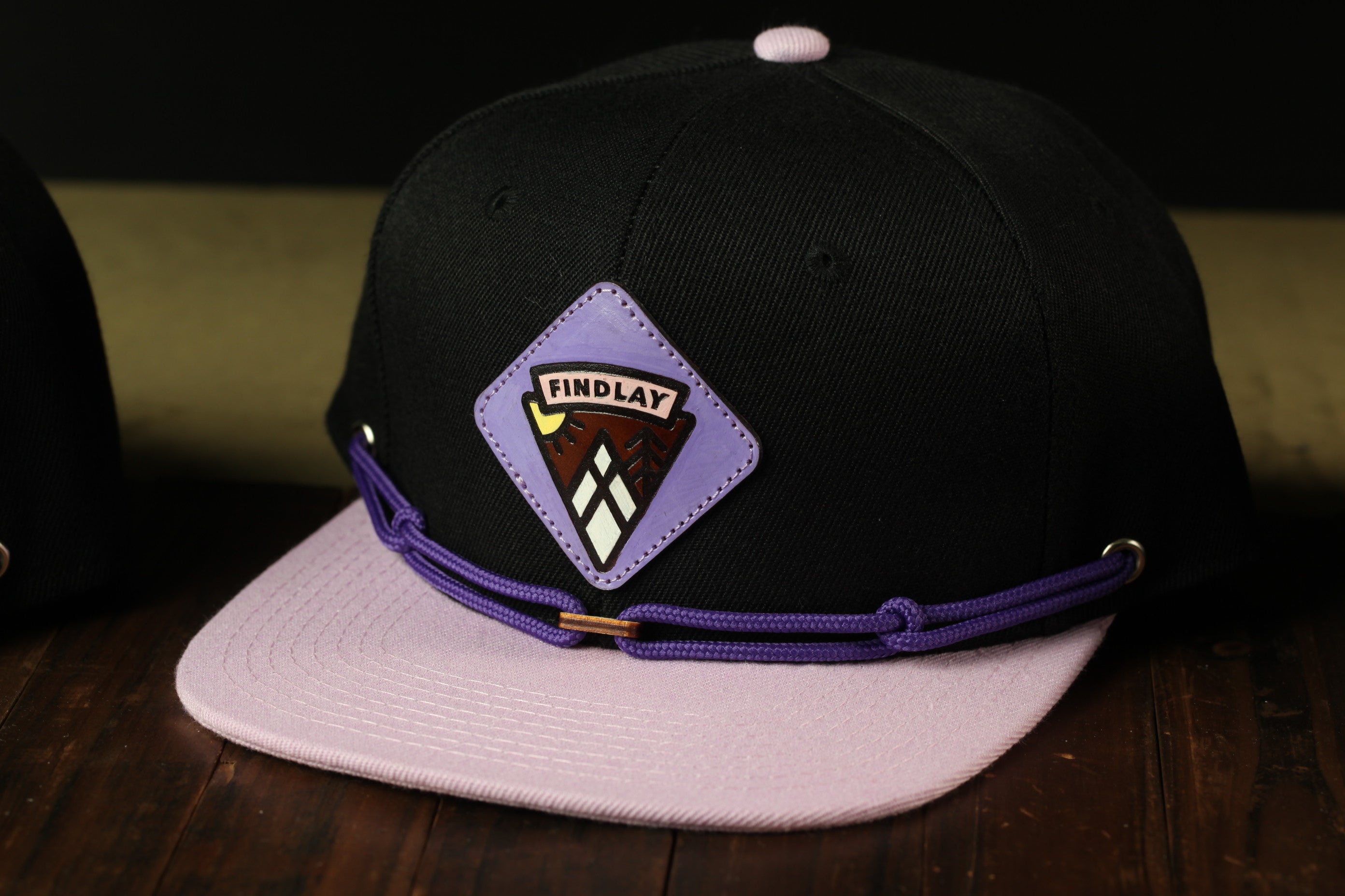 A black baseball cap with a purple band and a purple and white logo on the front, sitting on a wooden surface.