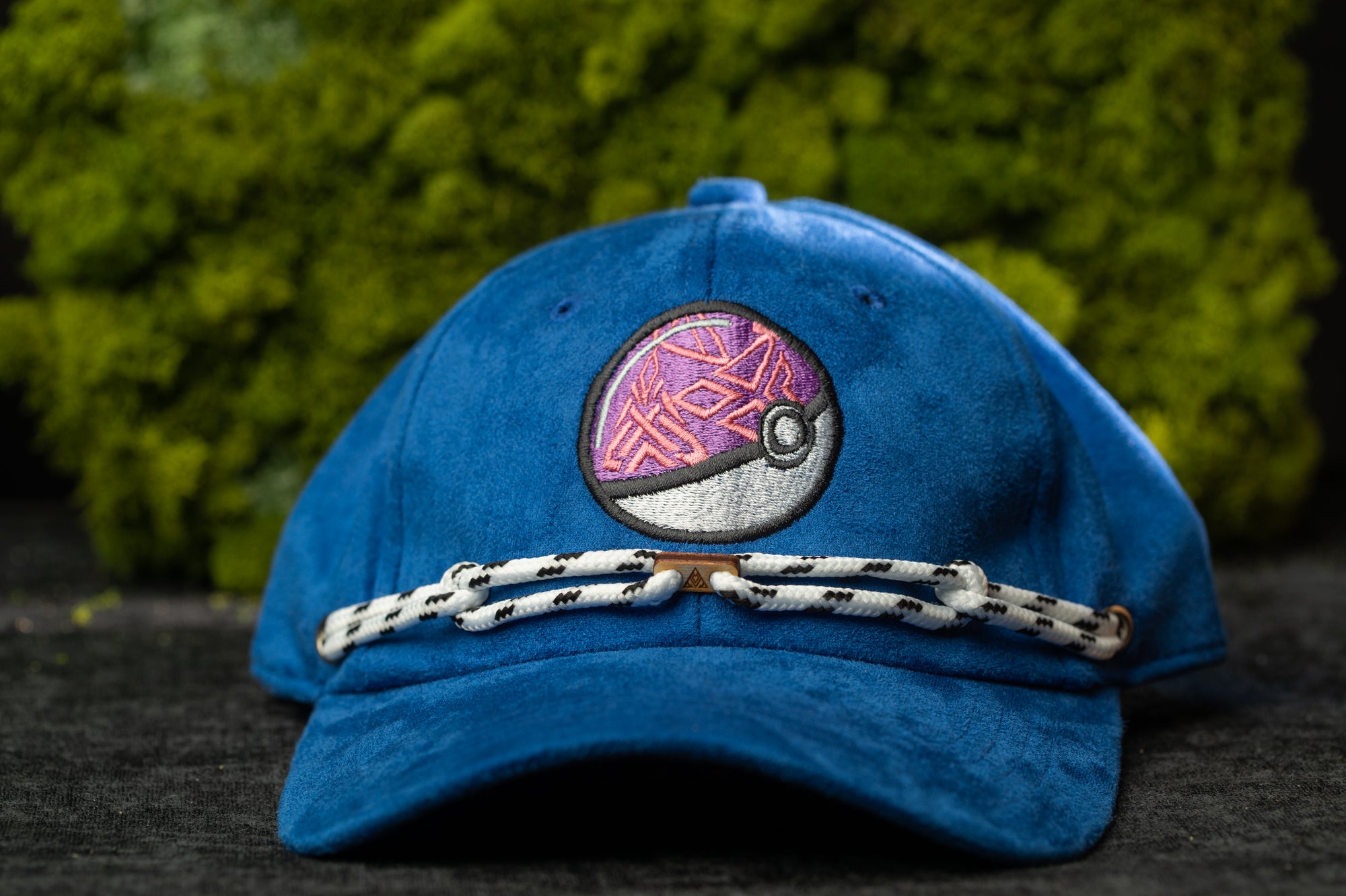 A blue baseball cap with a Pokemon-themed patch on the front, placed on a dark surface against a background of green moss.