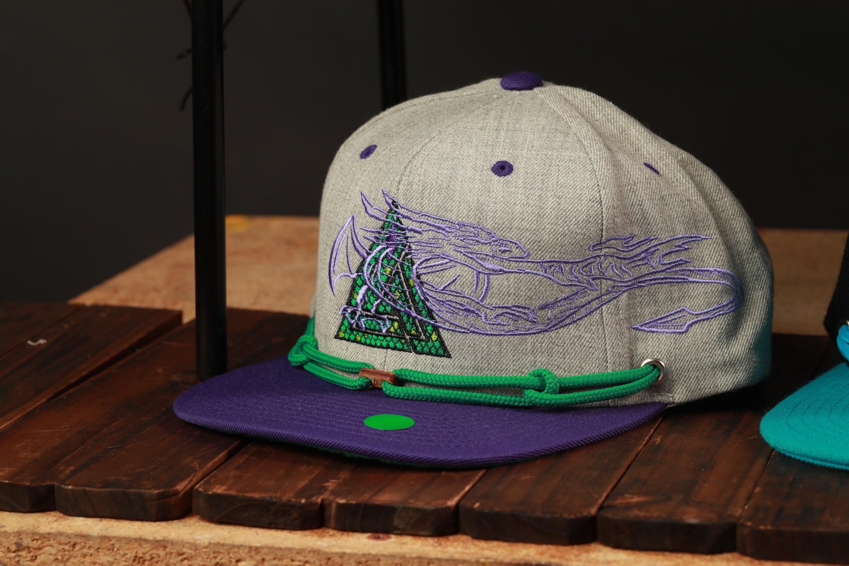 A gray baseball cap with purple and green accents, featuring a graphic design on the front, is displayed on a wooden surface.