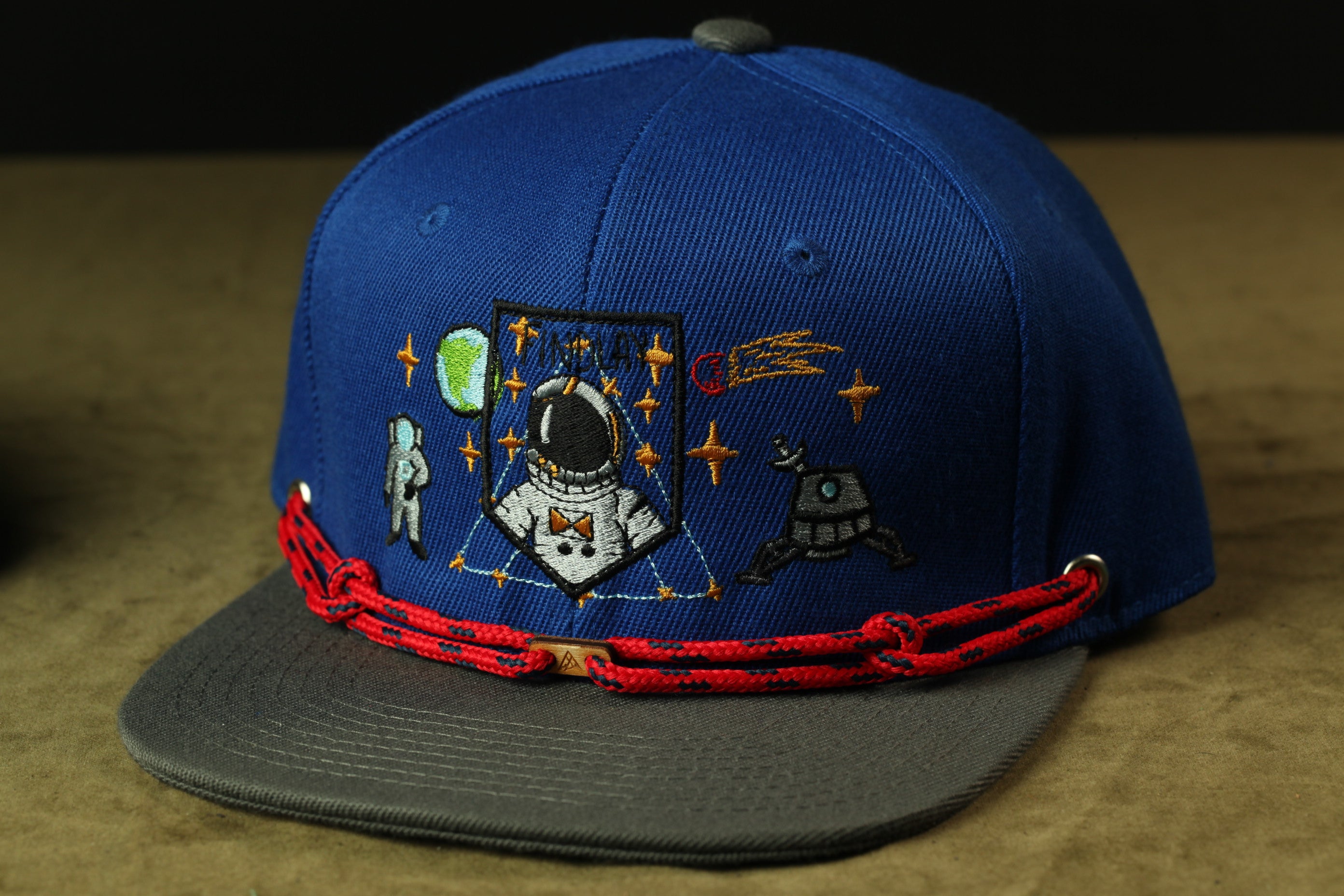 A blue baseball cap with various space-themed embroidered designs, including a space suit, planets, and stars.
