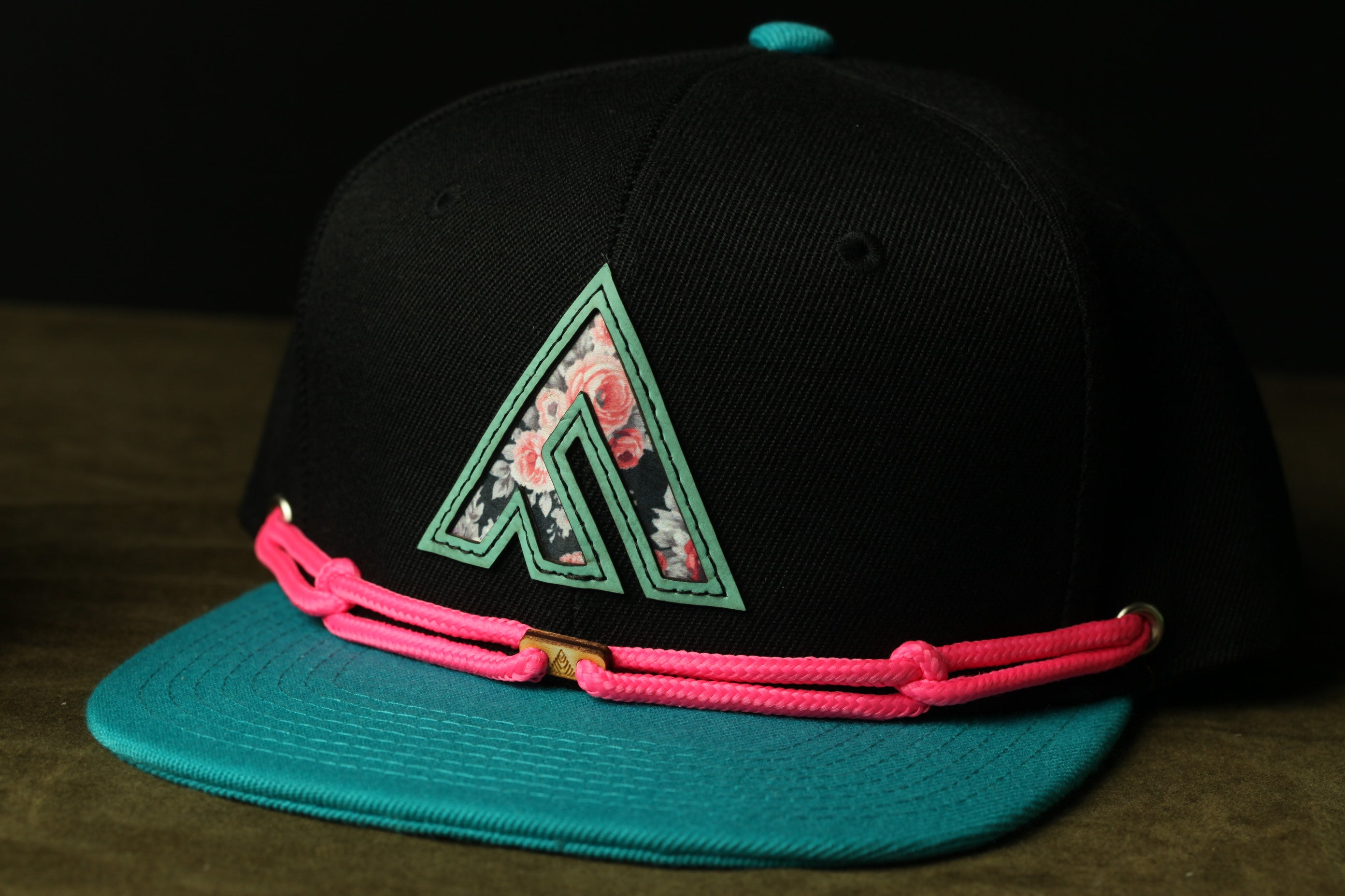 A black baseball cap with a floral pattern and a pink braided strap.