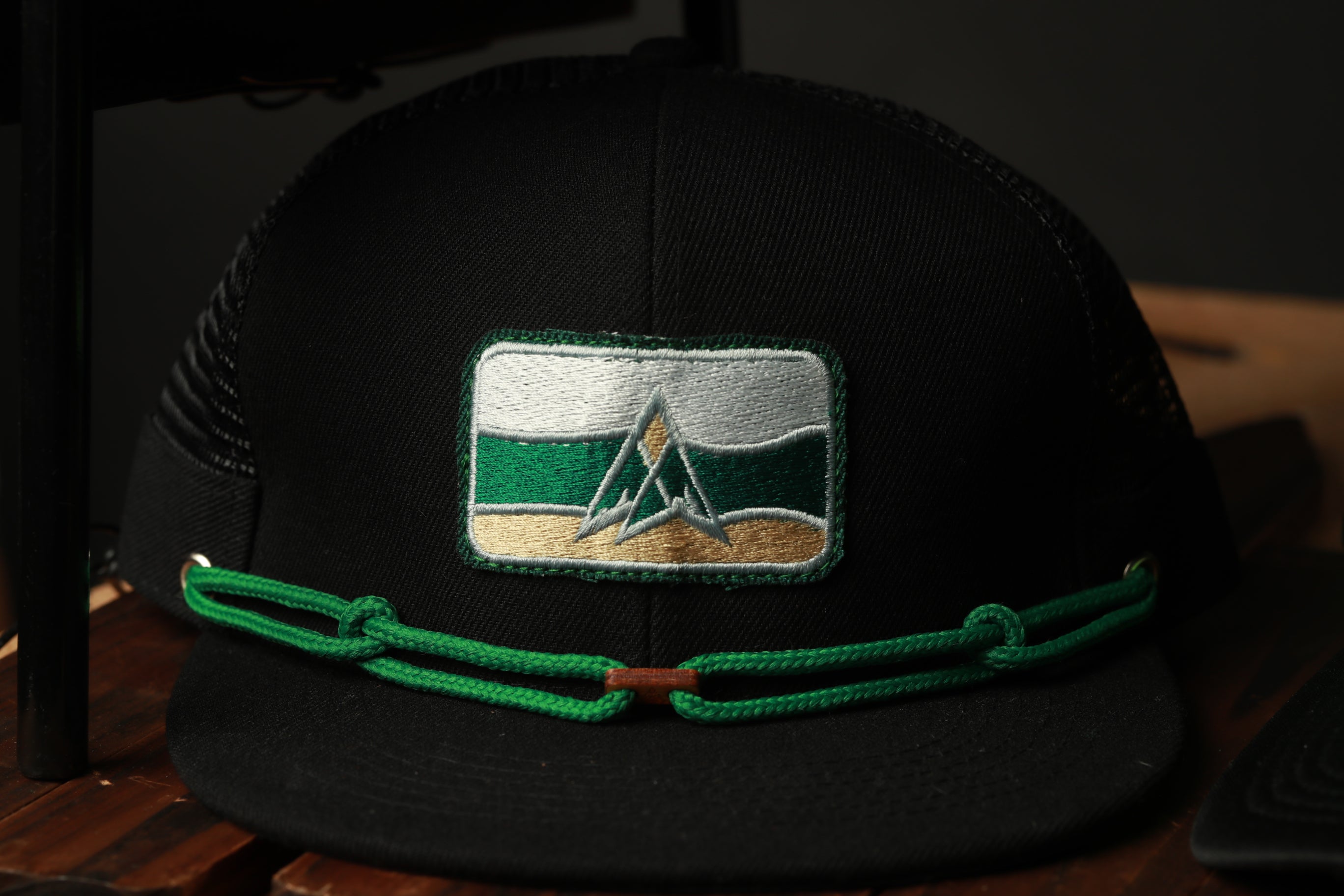 A black baseball cap with a green and white patch featuring a mountain design, and a green chain strap.