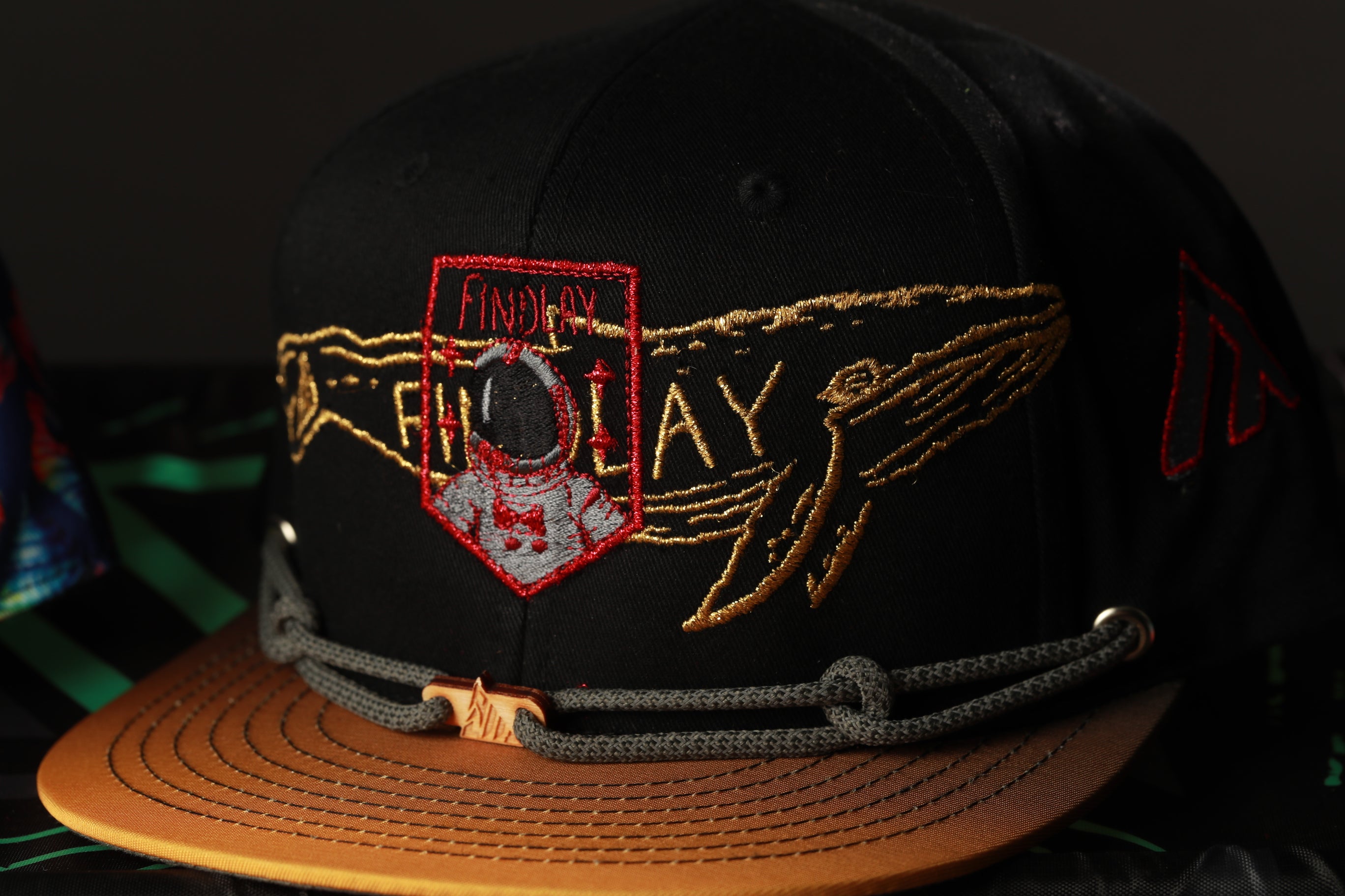 A black baseball cap with gold embroidered designs, including a shield-shaped emblem and a whale-like figure.