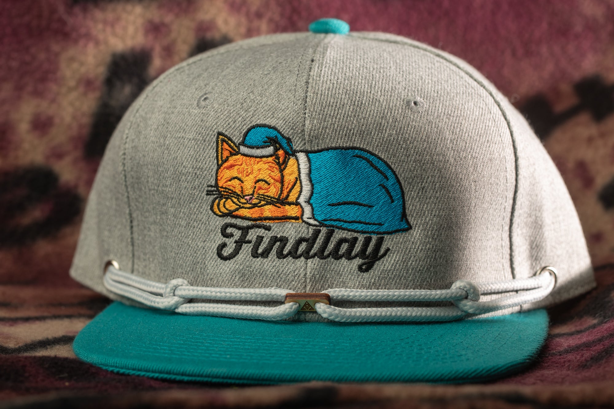A gray baseball cap with a teal brim and a blue cat embroidered on it, along with the word "Findlay" printed on it.