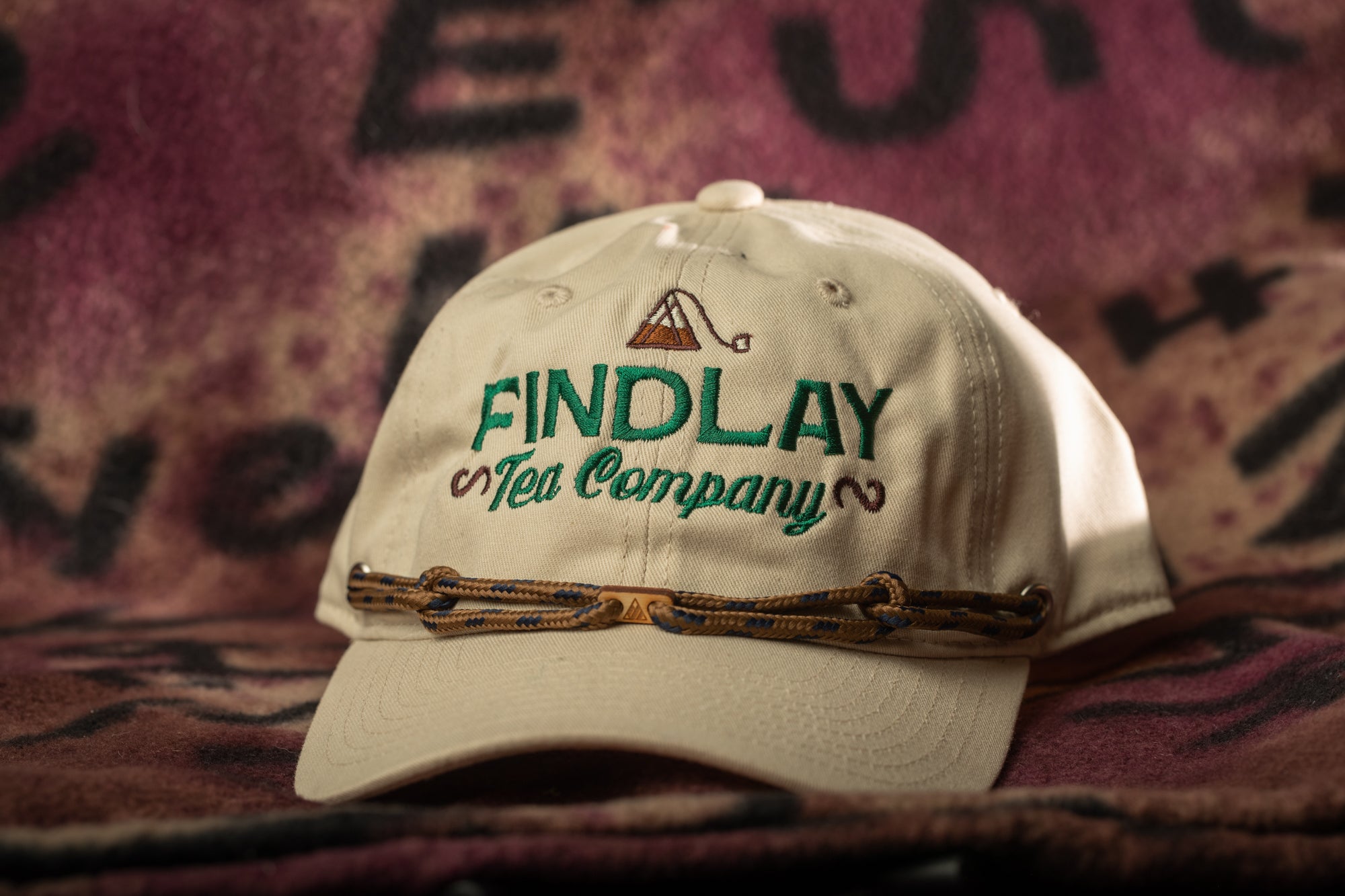 A beige baseball cap with the text "FIND LAY Tea Company" printed on it, sitting on a patterned fabric background.
