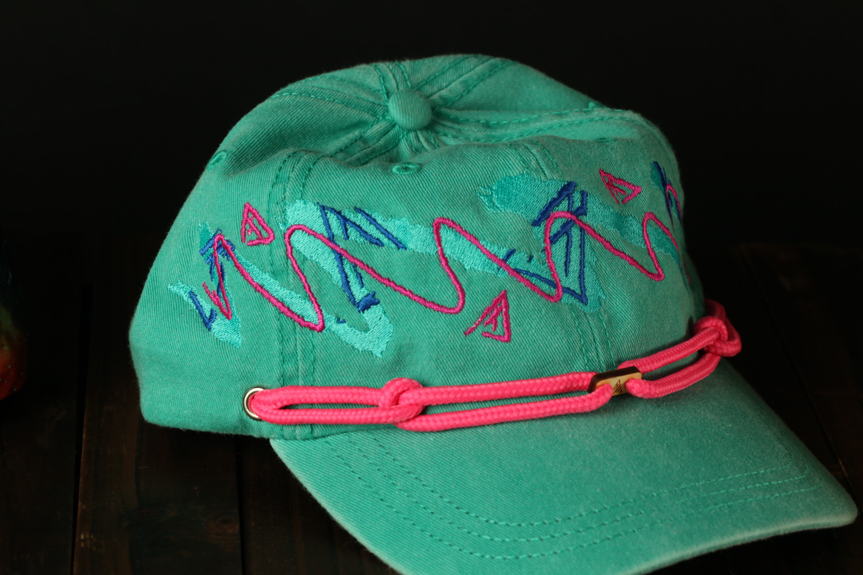 A turquoise baseball cap with pink braided straps and colorful embroidered designs on the front.