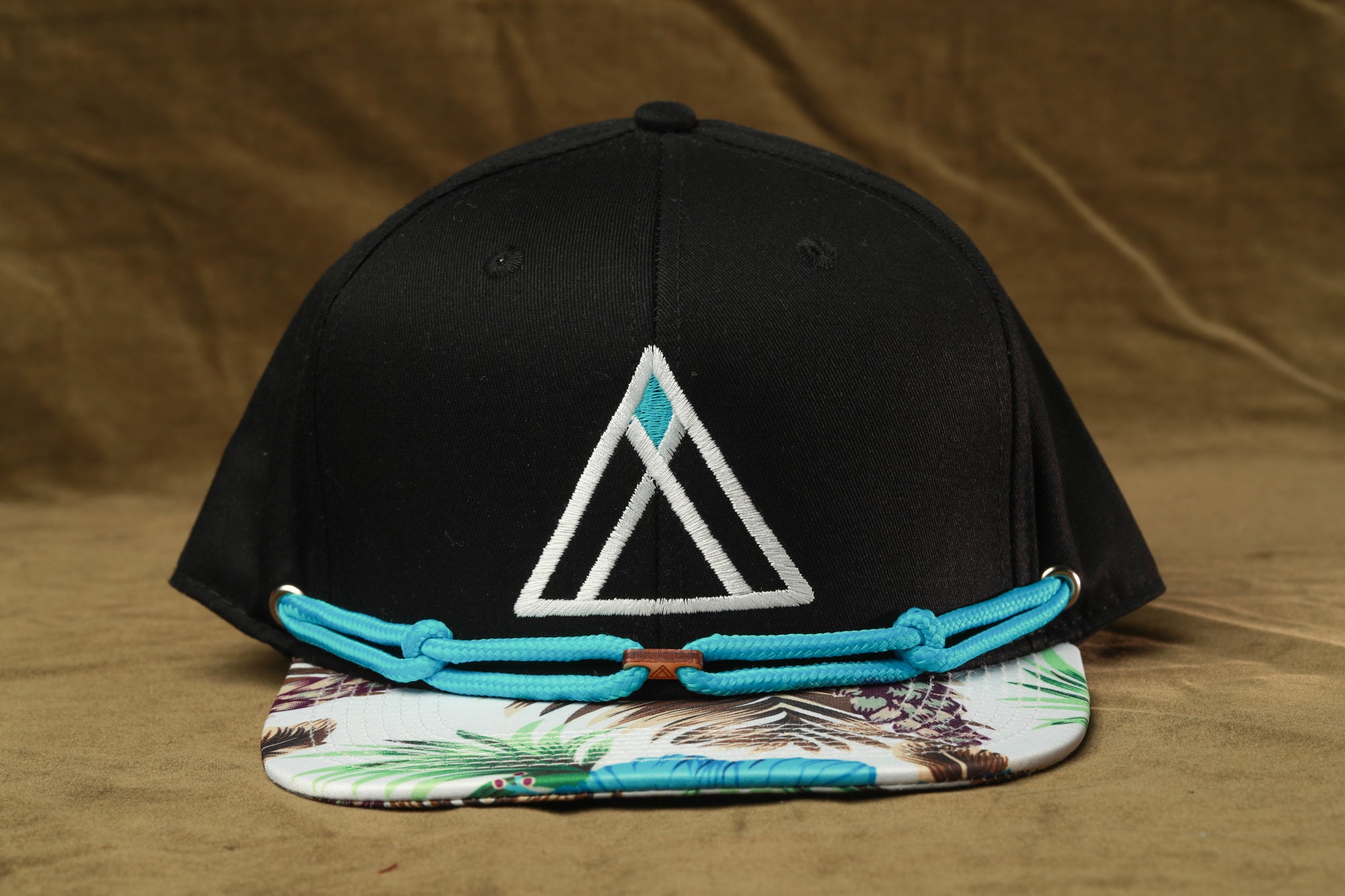 A black baseball cap with a white triangle logo and a colorful printed pattern on the brim, resting on a brown surface.