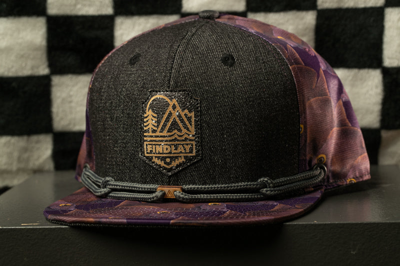A black baseball cap with a floral pattern and a gold-colored logo on the front, placed on a black surface against a checkered background.