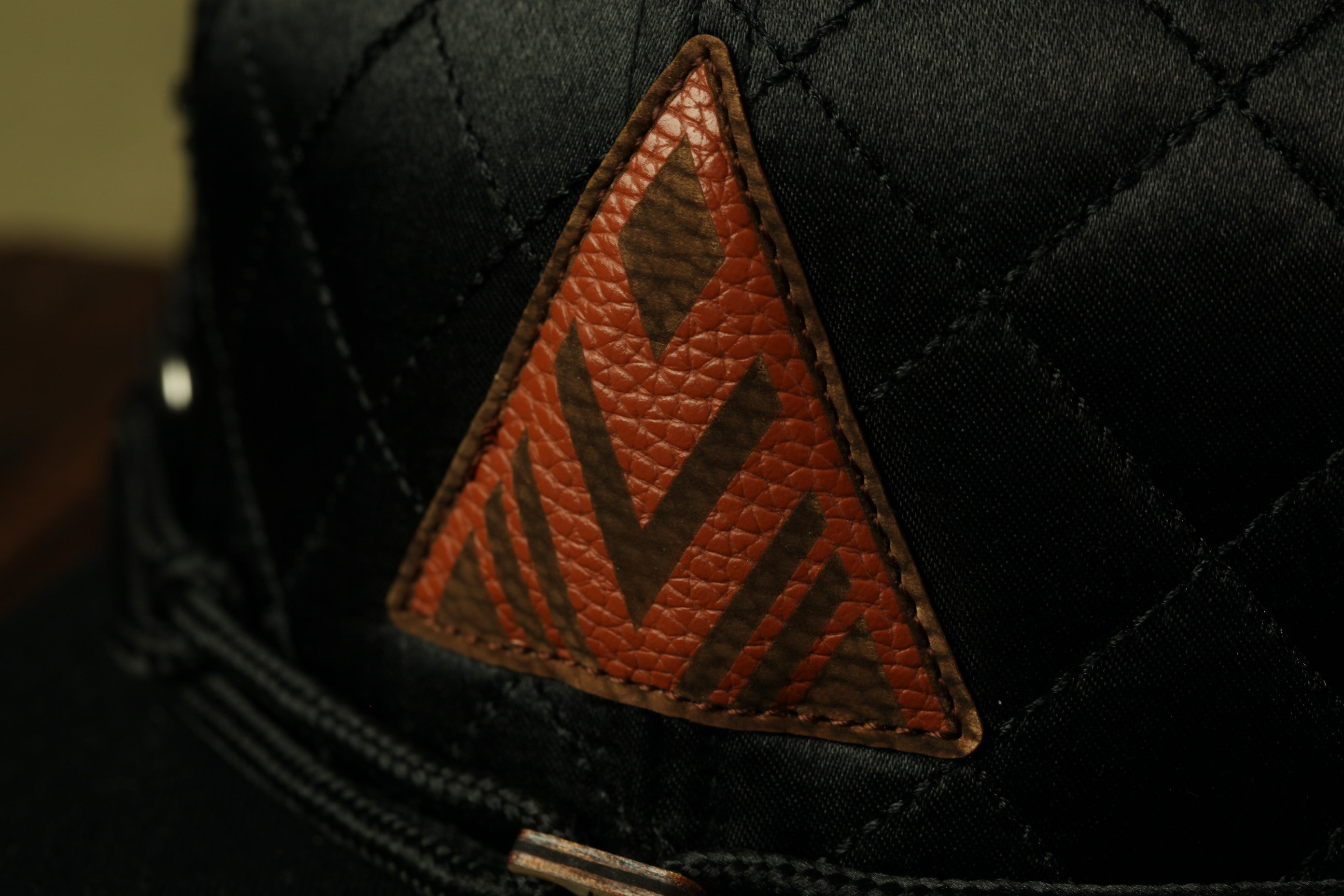 A close-up view of a black baseball cap with a distinctive triangular patch featuring an orange and brown geometric pattern.