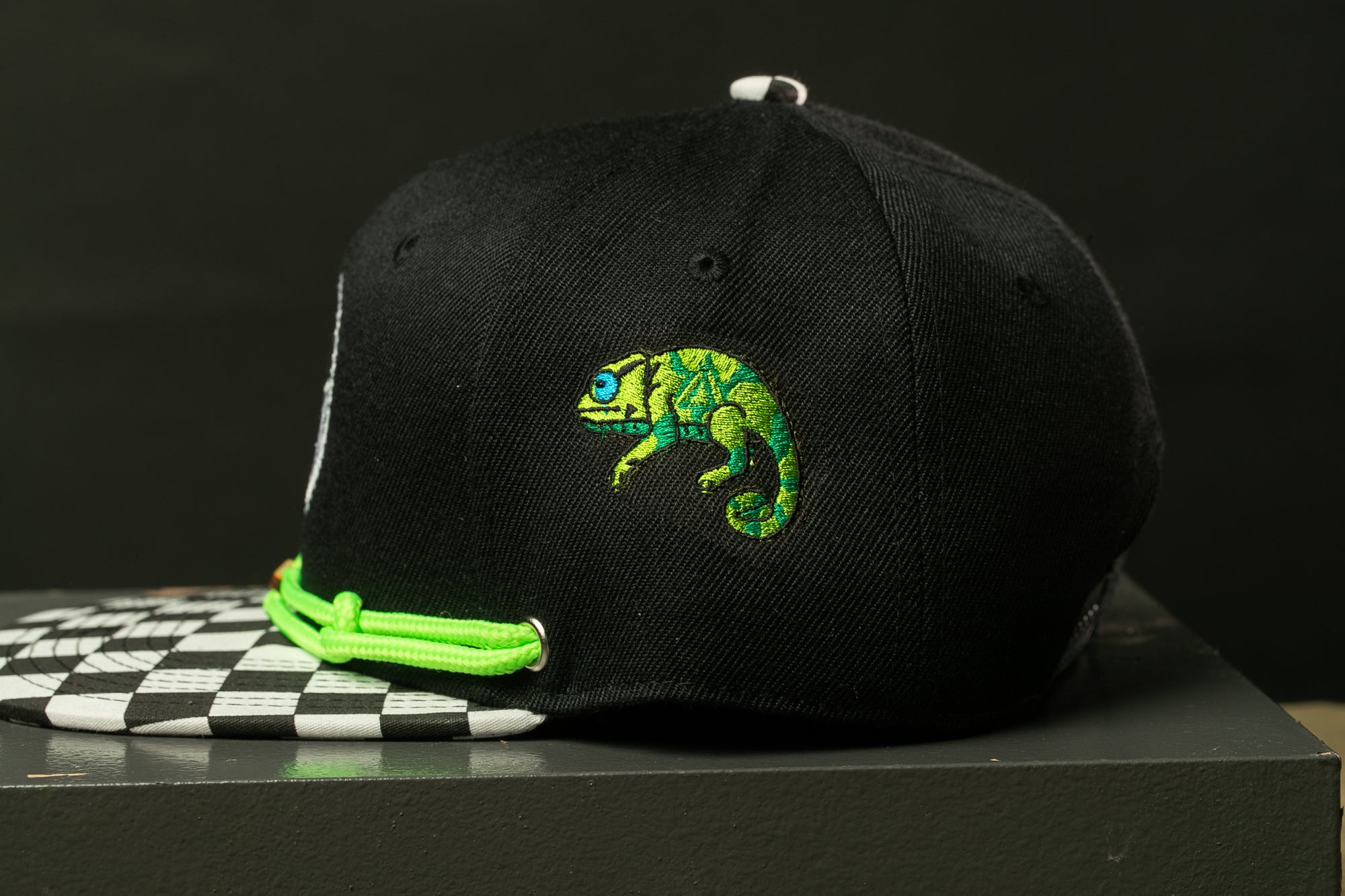 A black baseball cap with a green and yellow chameleon embroidered on it, sitting on a black and white checkered surface.