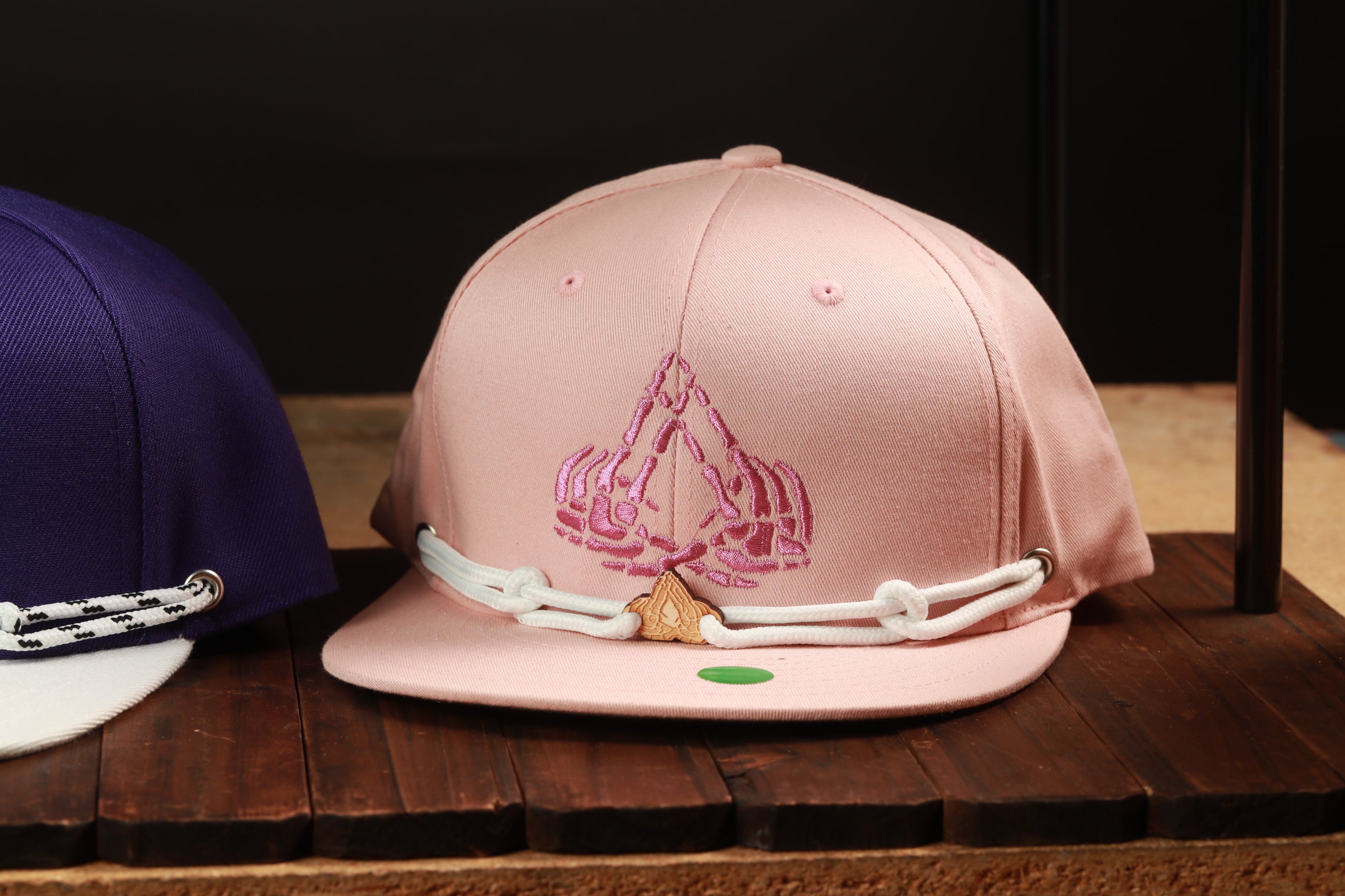 A pink baseball cap with a pink and white embroidered design on the front, sitting on a wooden surface.
