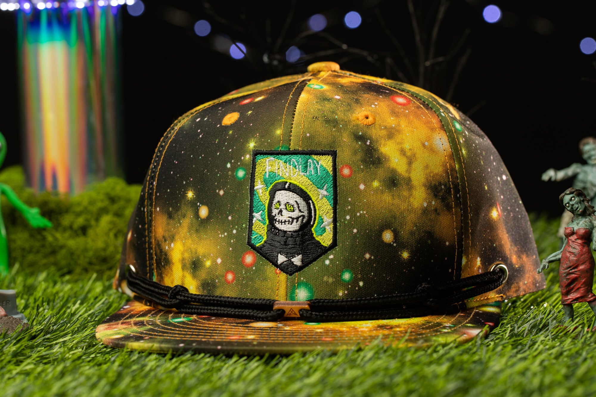 A colorful, space-themed baseball cap with a skull and crossbones design, placed on a grassy surface with various small figurines and a colorful, abstract background.