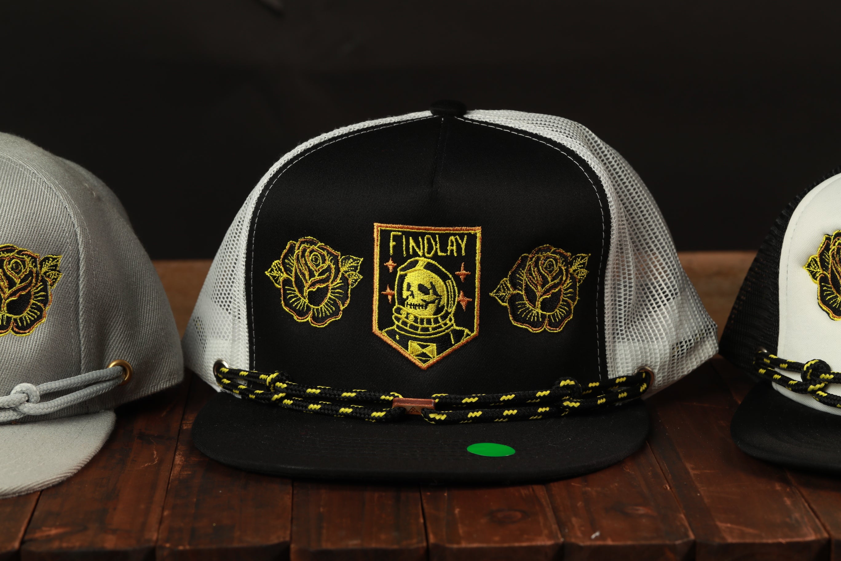 A collection of three baseball caps with unique designs, including a black and white cap with a skull and rose motif, a black and white cap with a skull and rose motif, and a gray cap with a skull and rose motif.