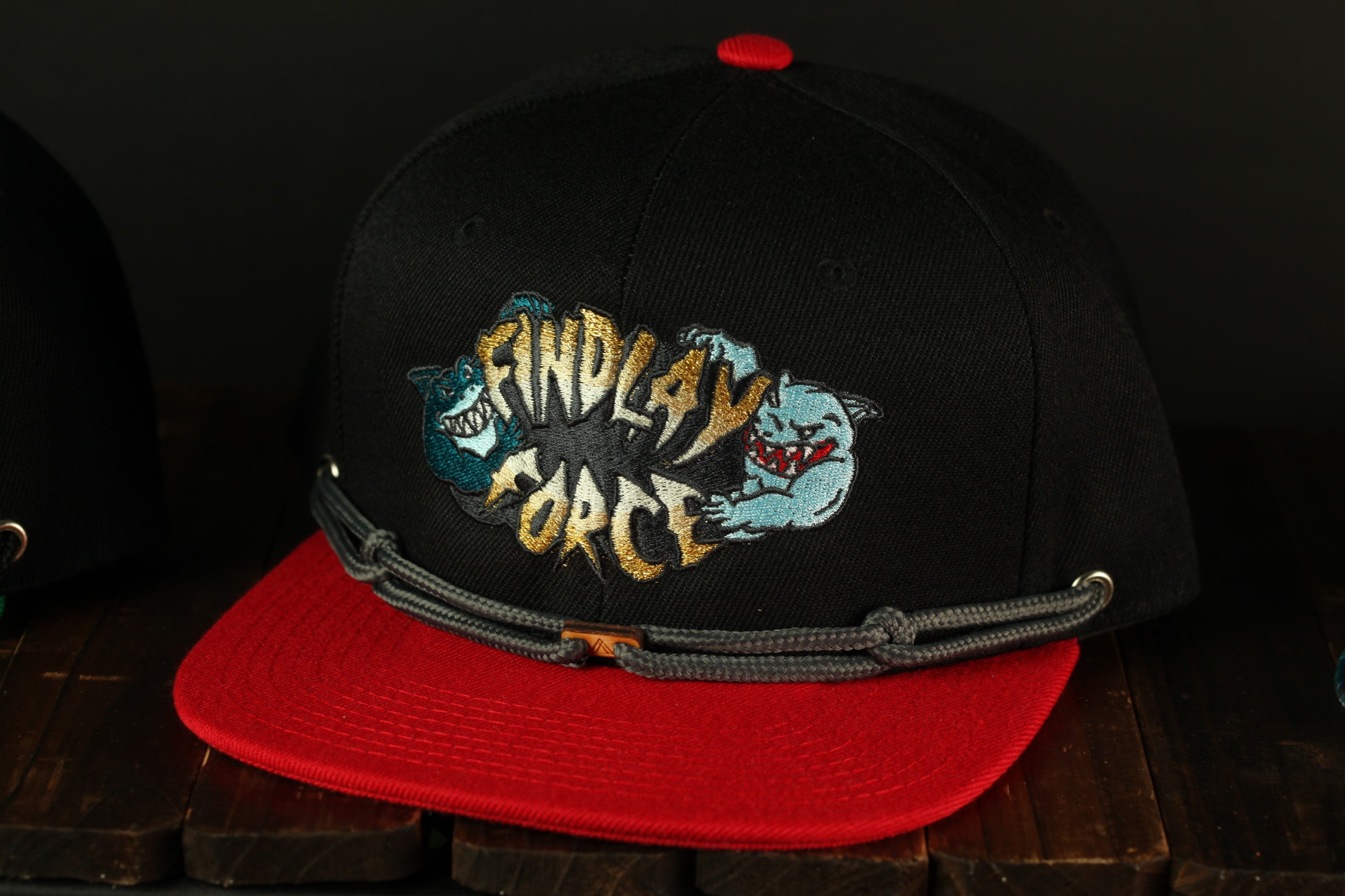 A black baseball cap with a red brim and a graphic design on the front, featuring a shark and the text "FINDlay Force".