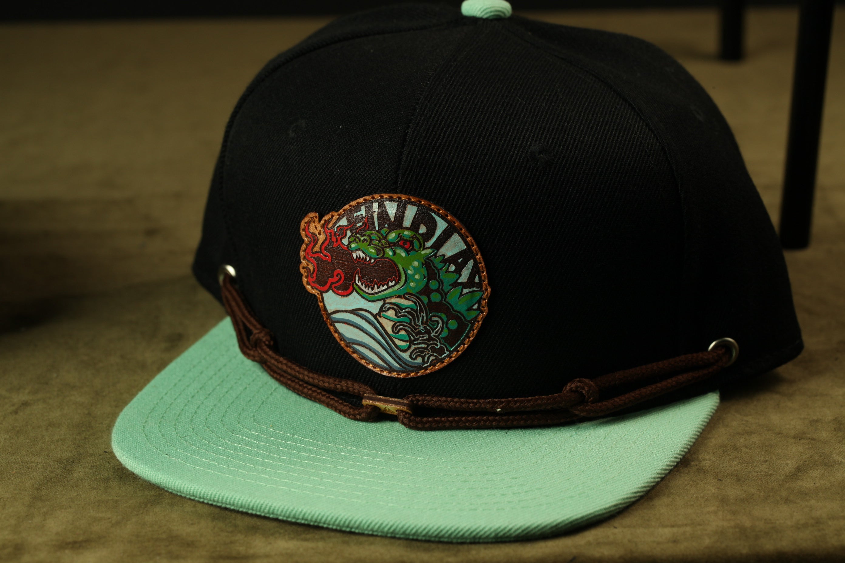 A black baseball cap with a green brim and a circular patch featuring a green and red design with a fish-like creature.