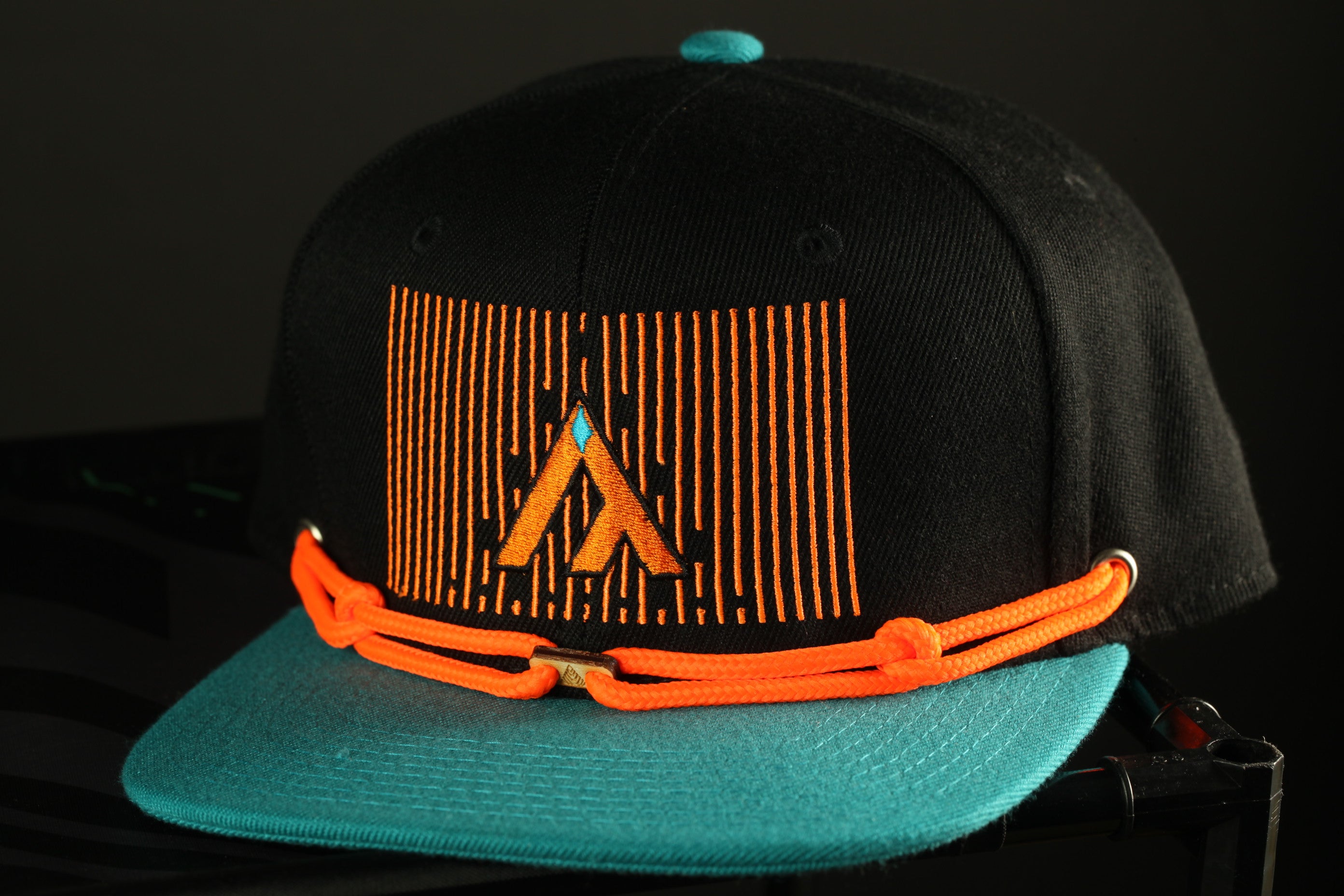 A black baseball cap with a teal brim and orange straps, featuring a graphic design on the front.