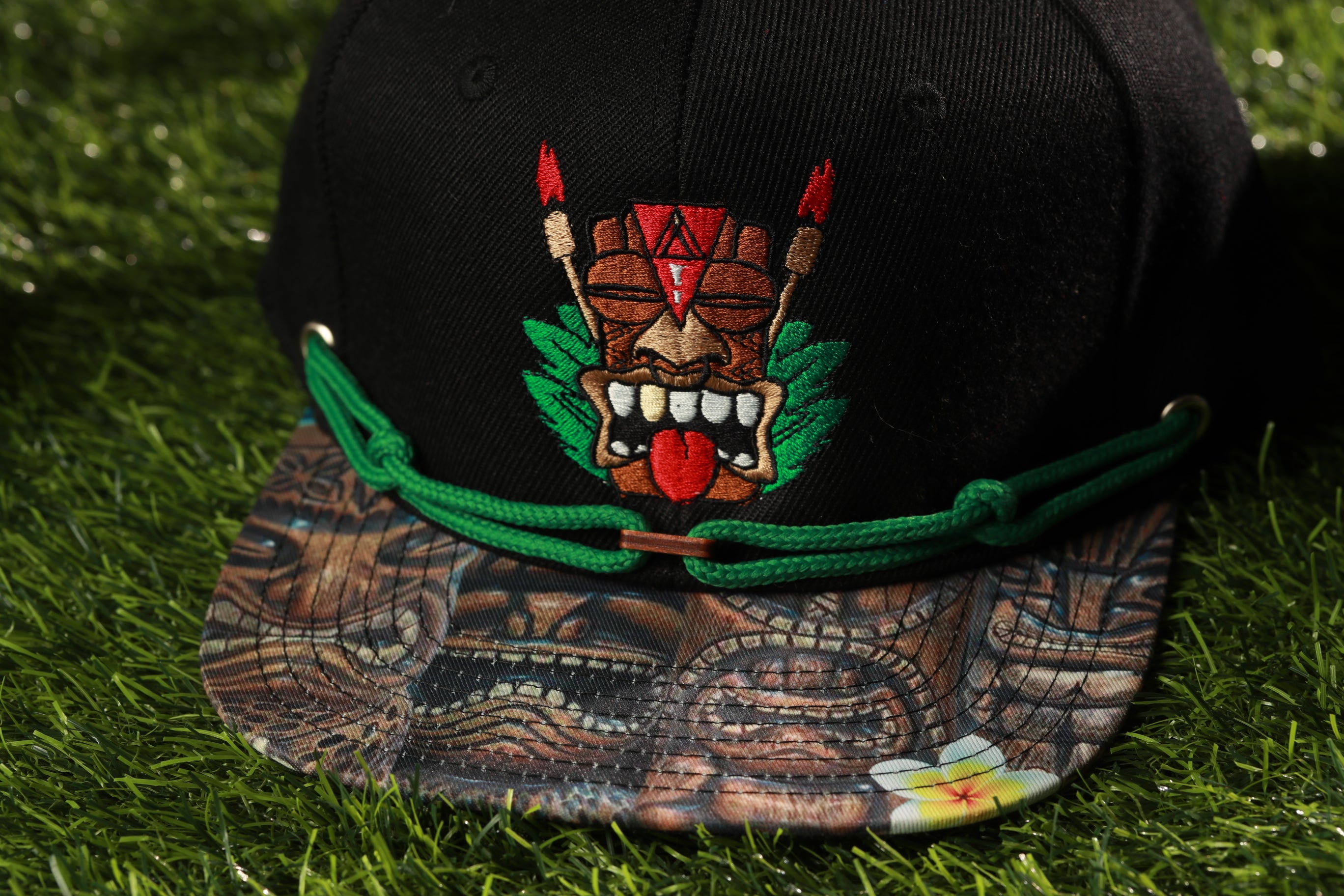 A black baseball cap with a colorful, tiki-style design on the front, including a mask and tropical foliage.