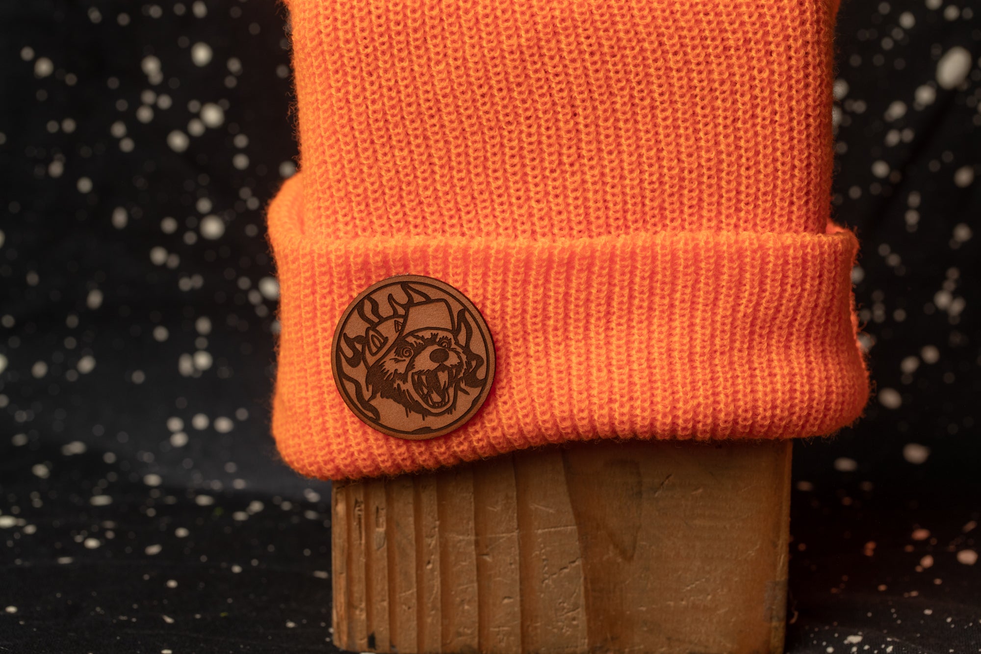 An orange knit cap with a circular badge featuring a wolf's face on it, placed on a wooden block against a dark background with white speckles.