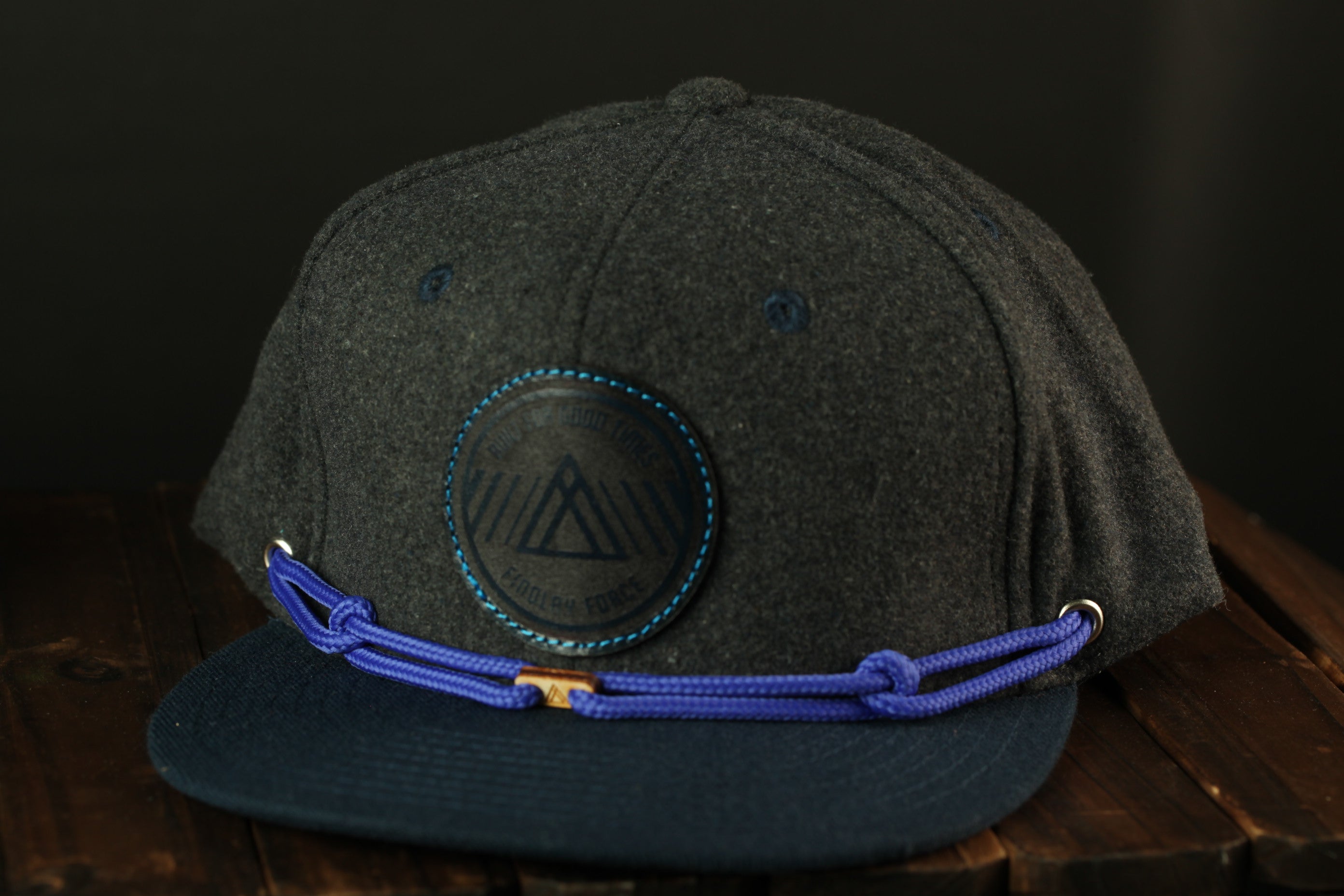 A dark grey baseball cap with a blue band and a circular logo on the front, placed on a wooden surface.