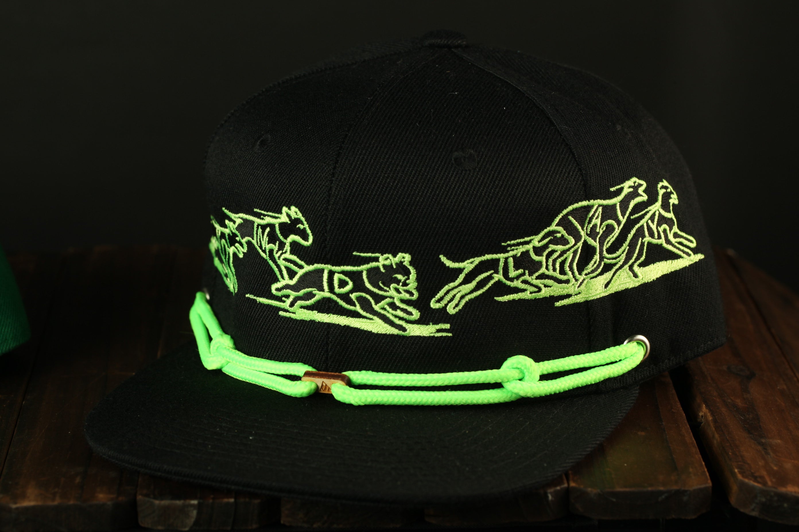 A black baseball cap with a green rope strap and a design of dogs running on a track.