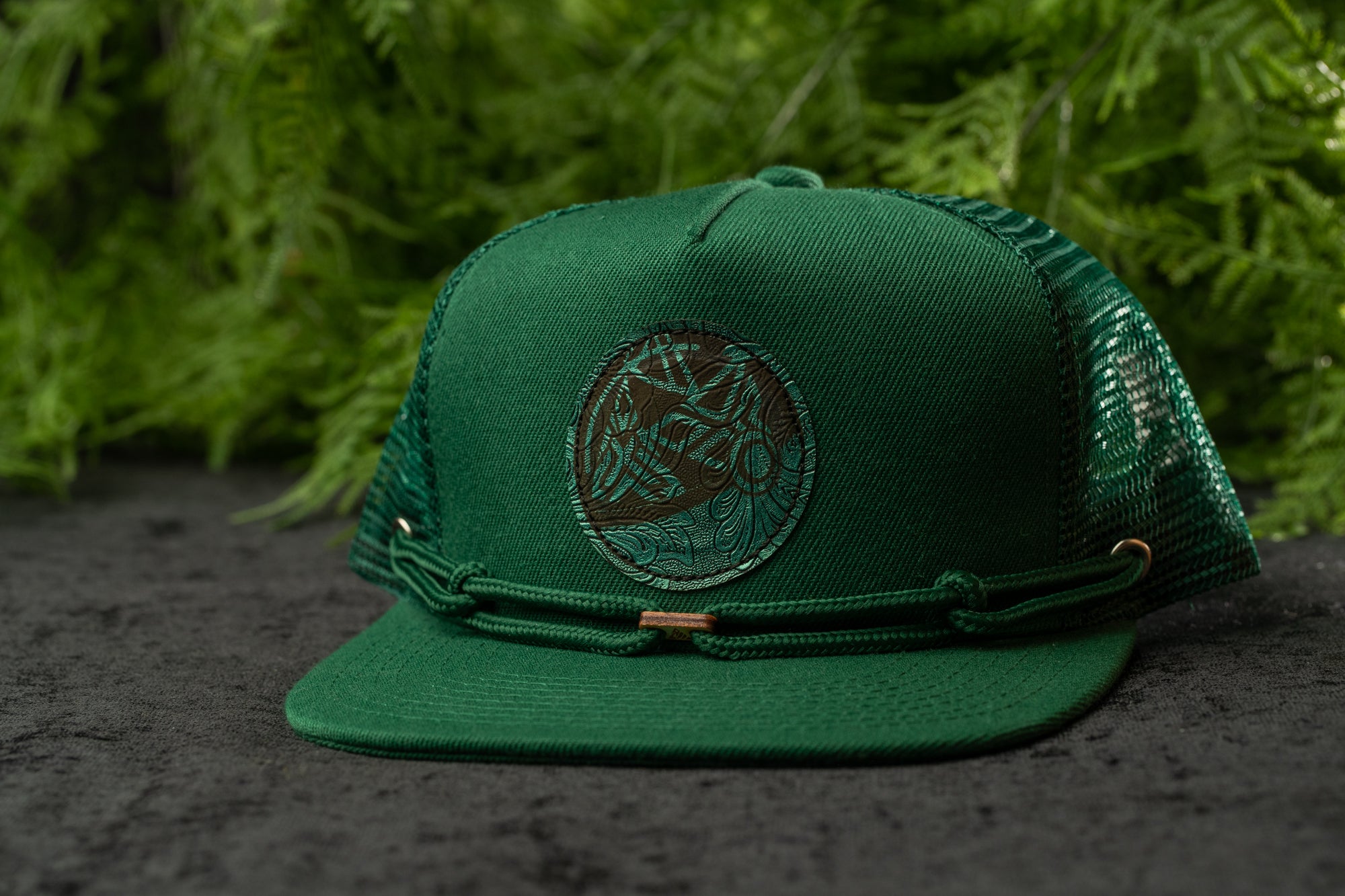 A green baseball cap with a circular logo on the front, placed on a surface with green foliage in the background.