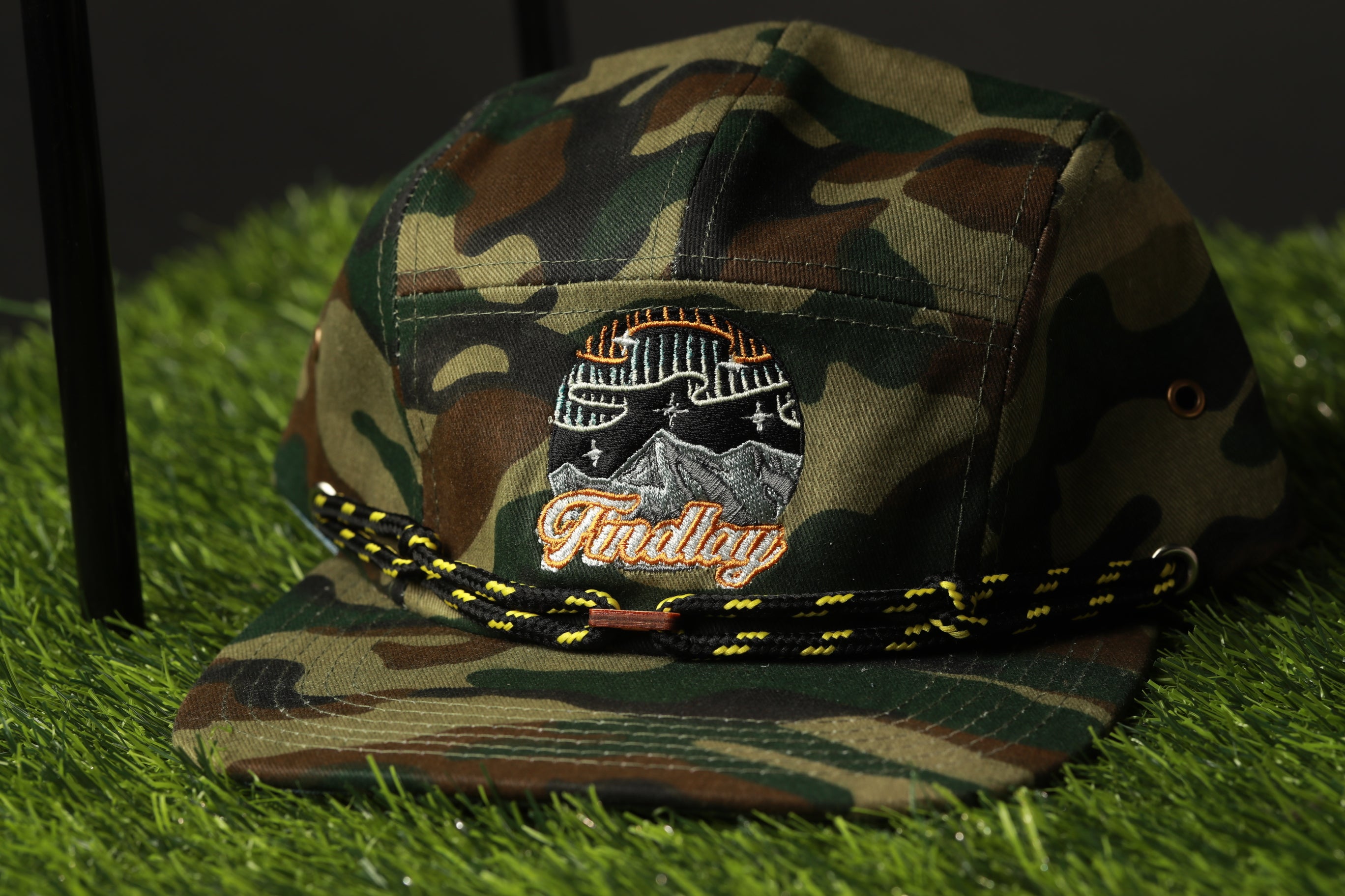 A camouflage baseball cap with a logo on it, placed on a grassy surface.