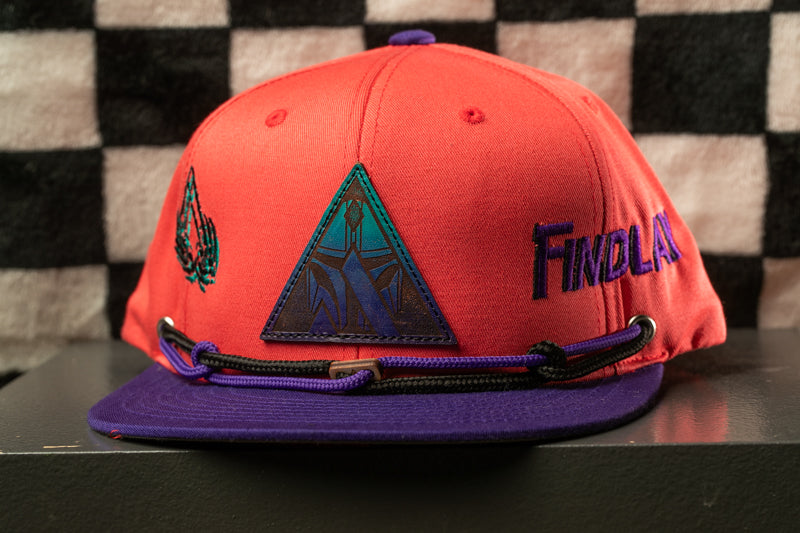 A red baseball cap with a purple brim and a black and purple strap, featuring a triangular patch with a graphic design on the front.