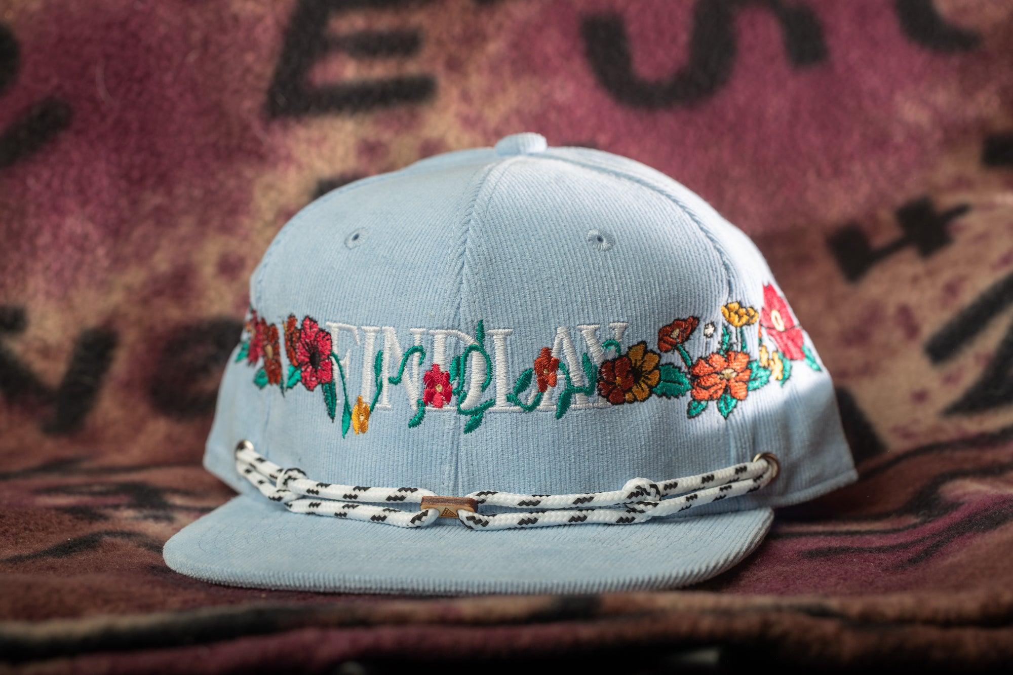 A light blue baseball cap with floral embroidery and a white lace strap is displayed against a colorful background.