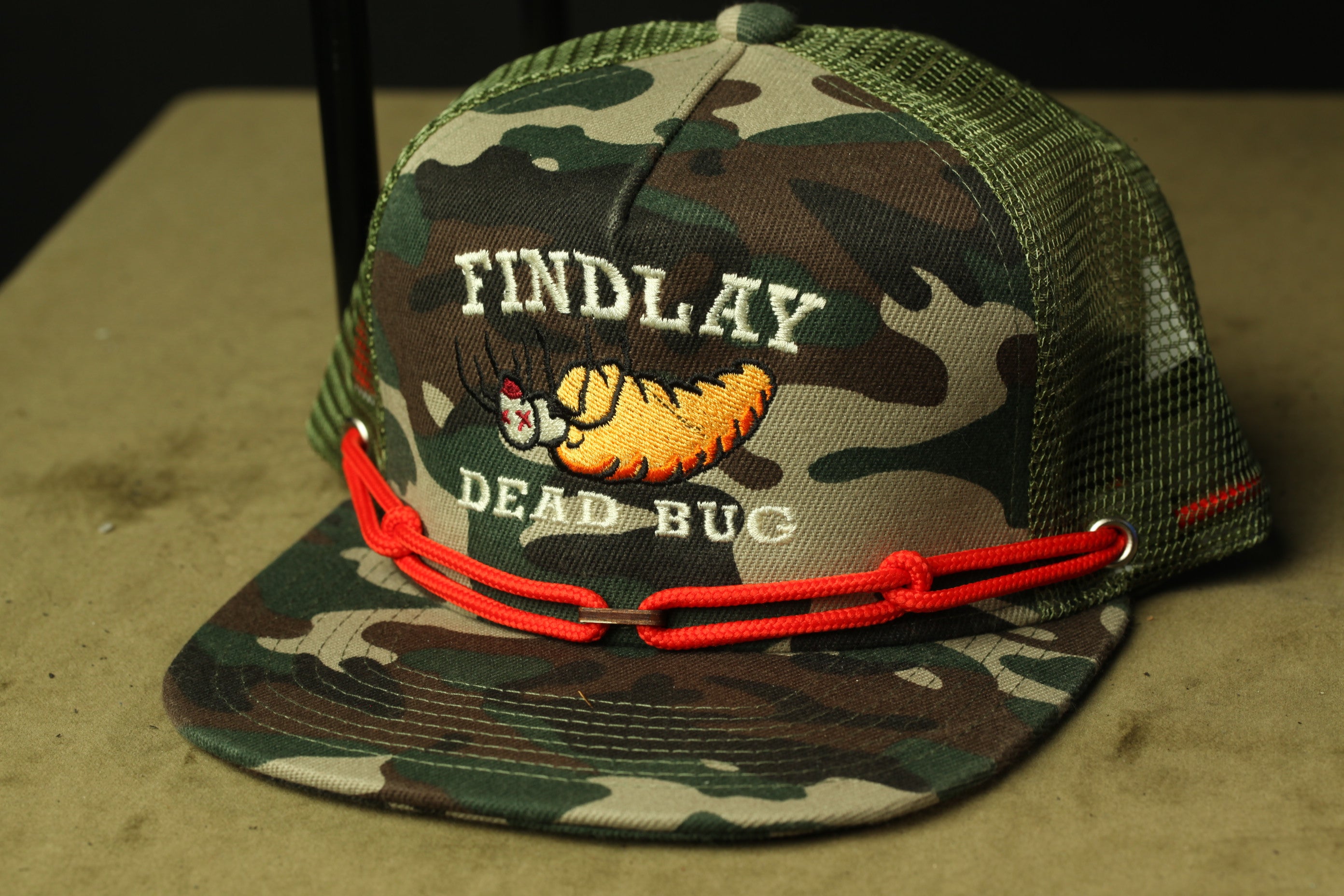A camouflage baseball cap with a red chain strap and a cartoon image of a dead bug on it.