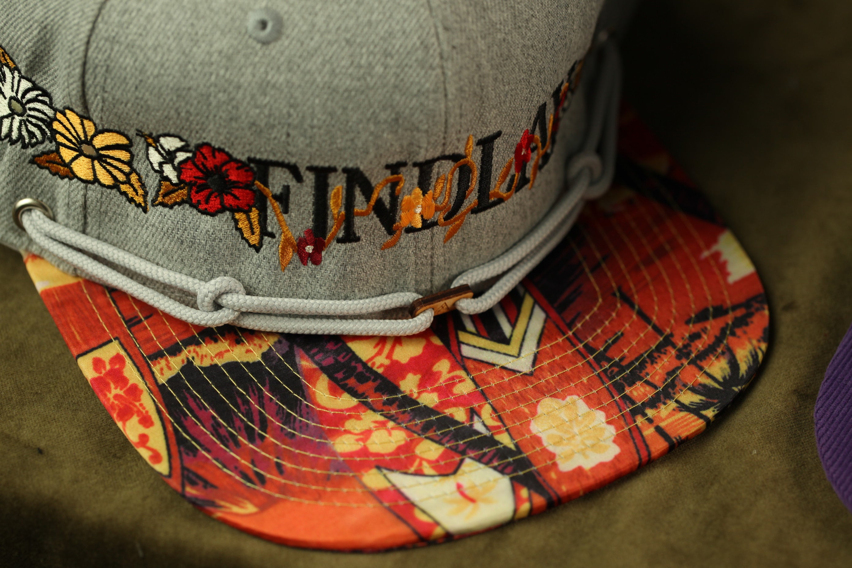A close-up image of a gray baseball cap with a colorful, patterned fabric and floral embroidery.
