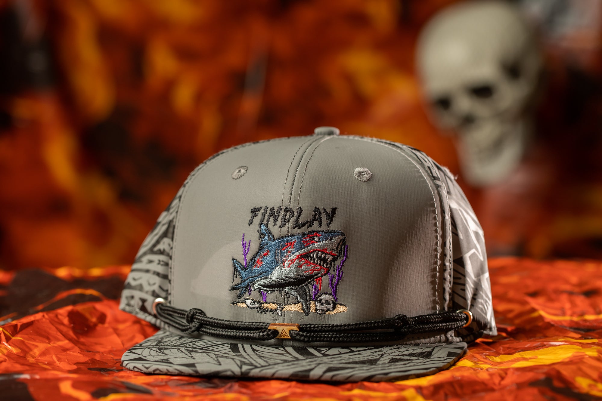 A gray baseball cap with a graphic of a shark and the text "FINDPLAY" printed on it, placed on a red and orange background.