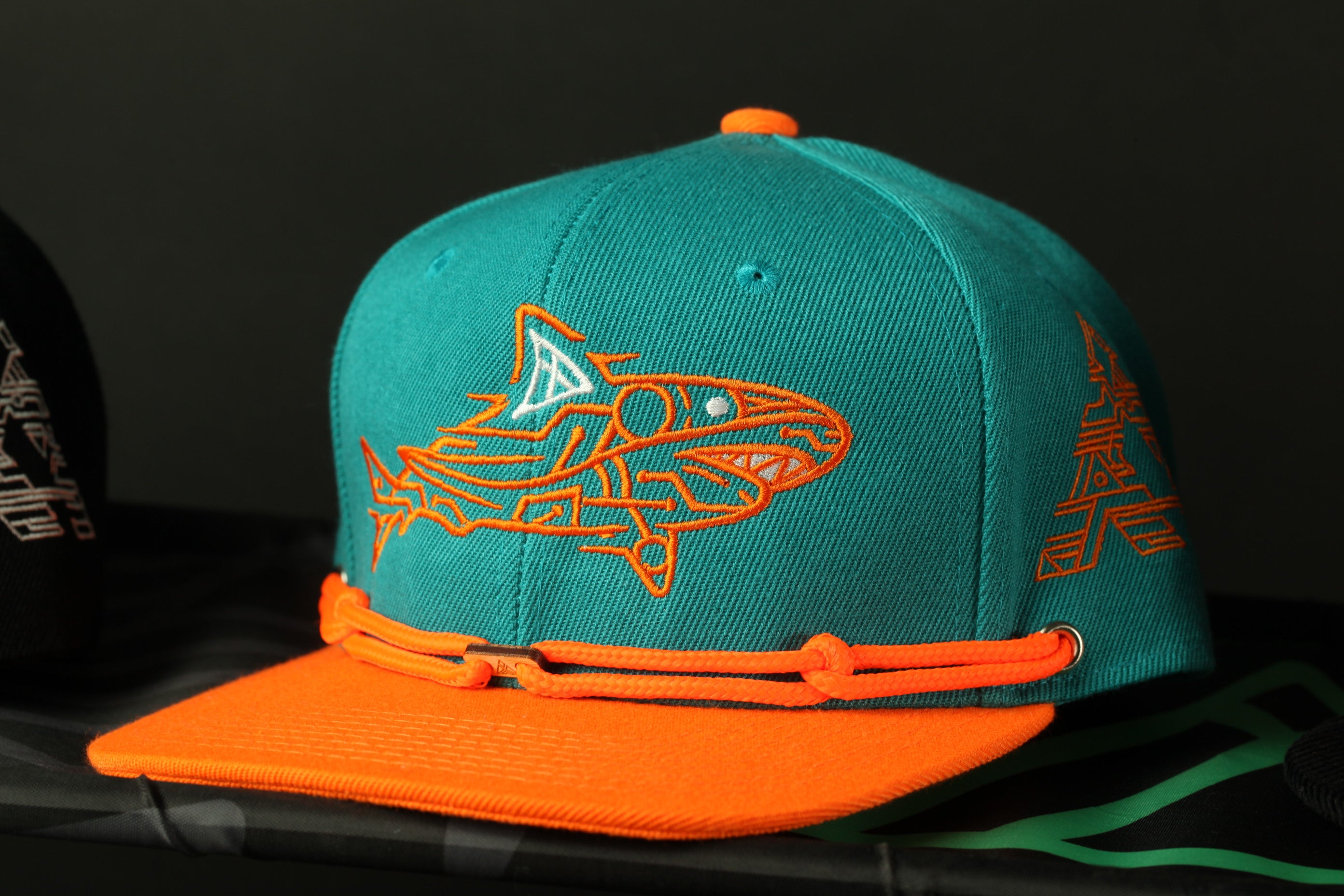 A turquoise baseball cap with an orange brim and a shark design on the front, displayed on a black surface.
