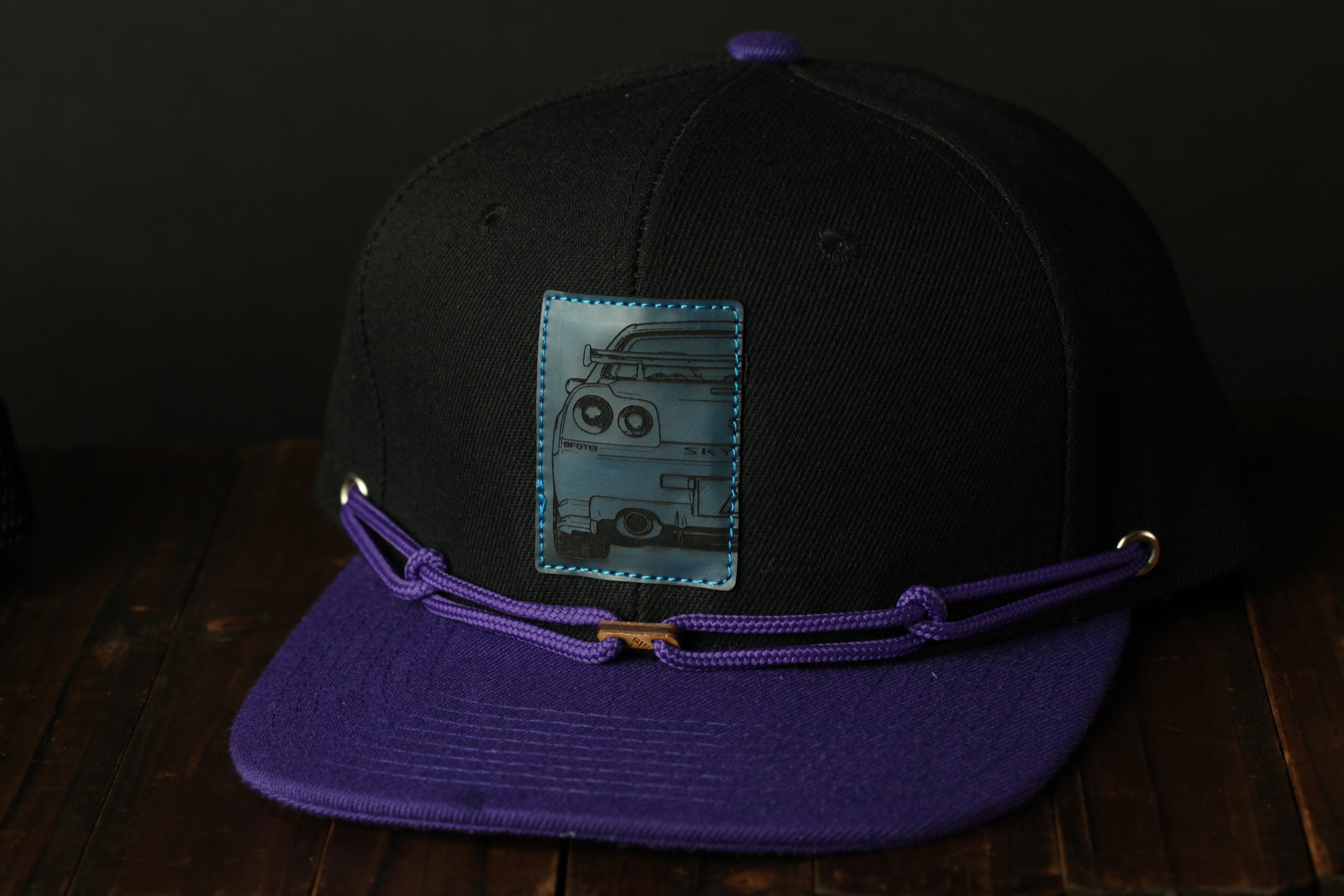 A black baseball cap with a purple brim and a blue patch featuring a cartoon image of a van.