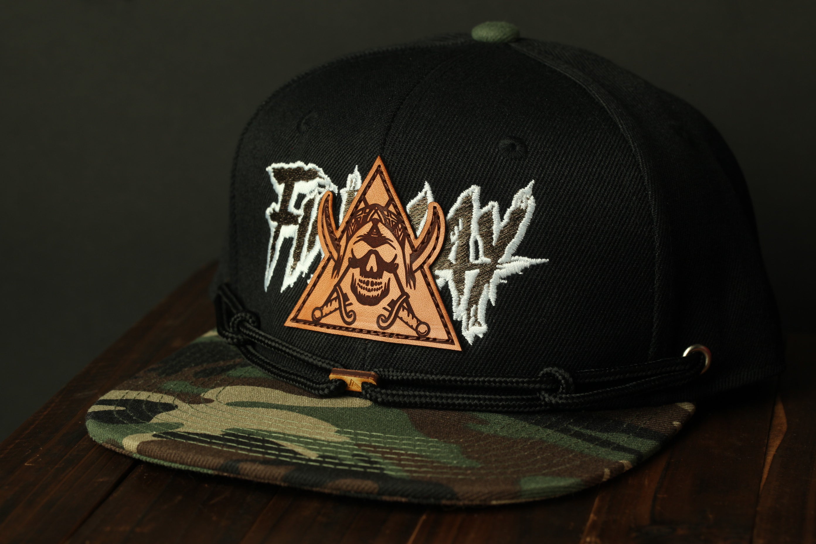 A black baseball cap with a camouflage pattern and a triangular patch featuring a skull and crossbones design.