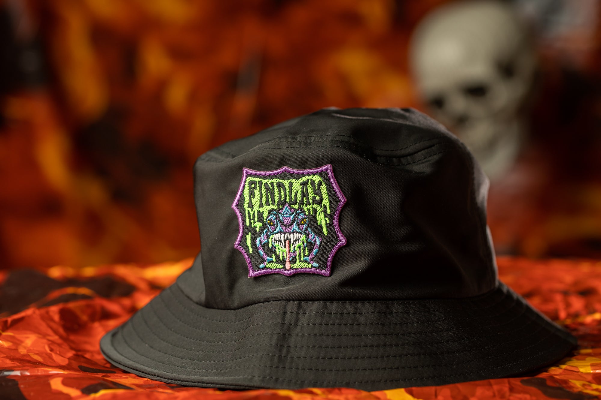 A black bucket hat with a colorful, graphic patch on the front, featuring a skull and a green, purple, and yellow design.