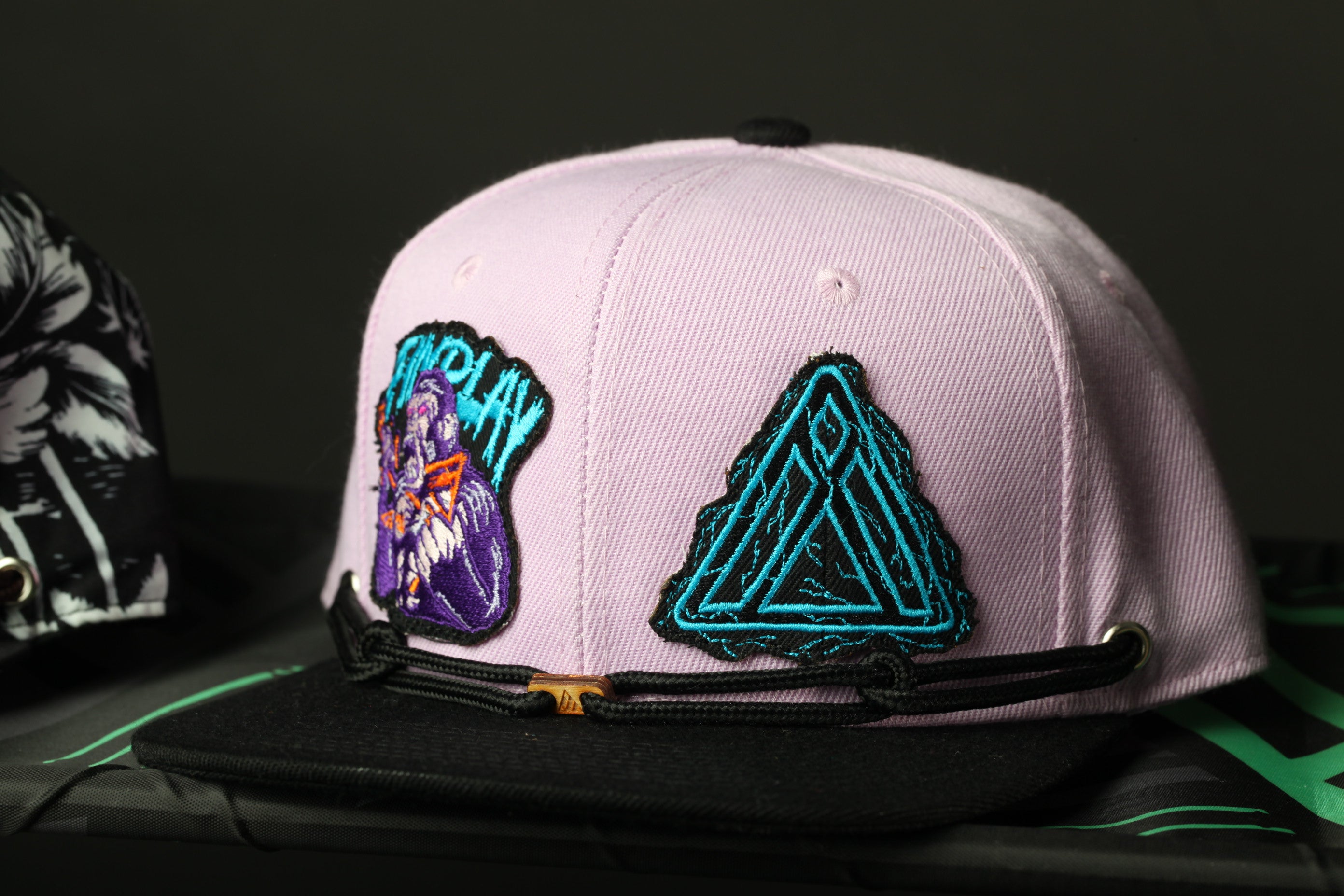 A pink baseball cap with two patches on it, one featuring a purple and blue design and the other a blue triangle design.