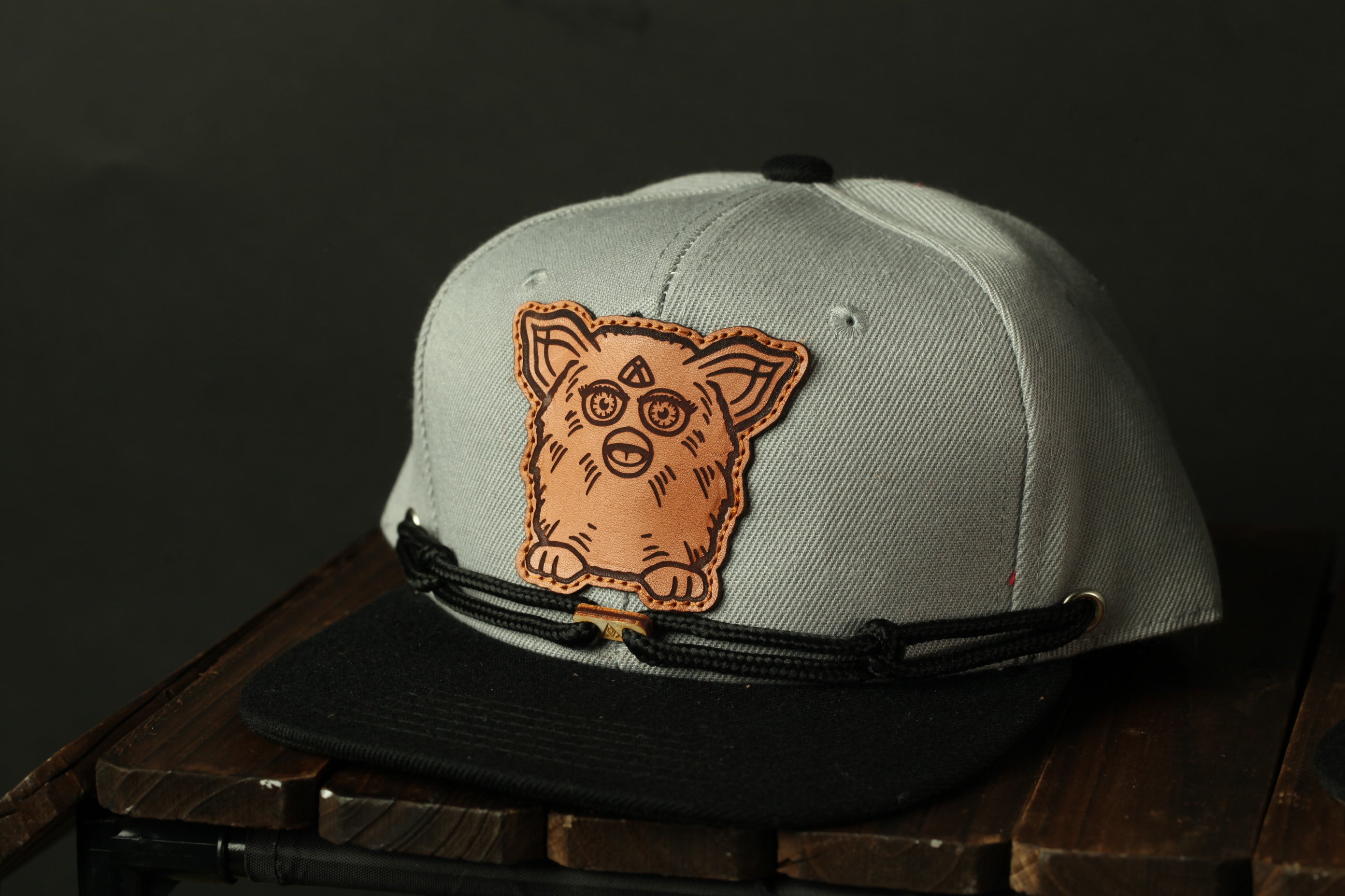 A gray baseball cap with a black brim and a brown embroidered character on the front, sitting on a wooden surface.
